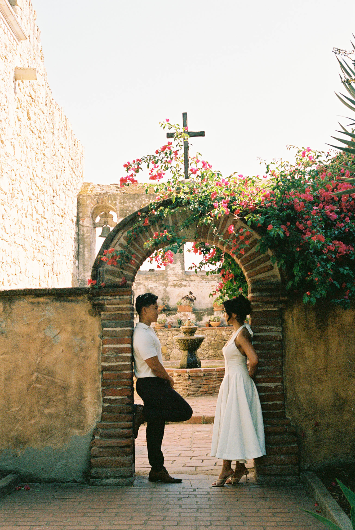 Couple standing under brick arch with bougainvillea at historic mission.