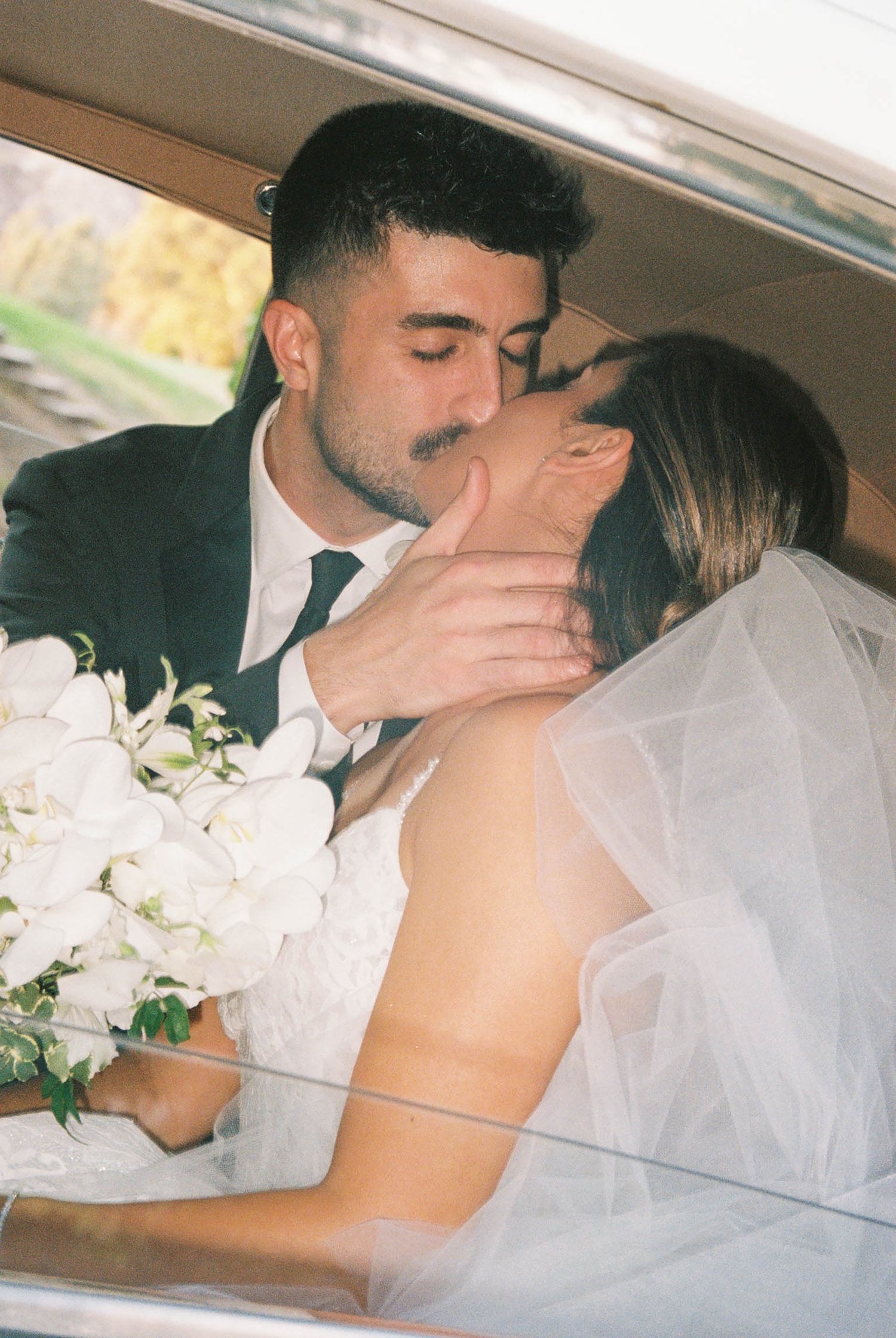 Bride and groom sharing a romantic kiss inside vintage car, bride holding white orchid bouquet with veil.