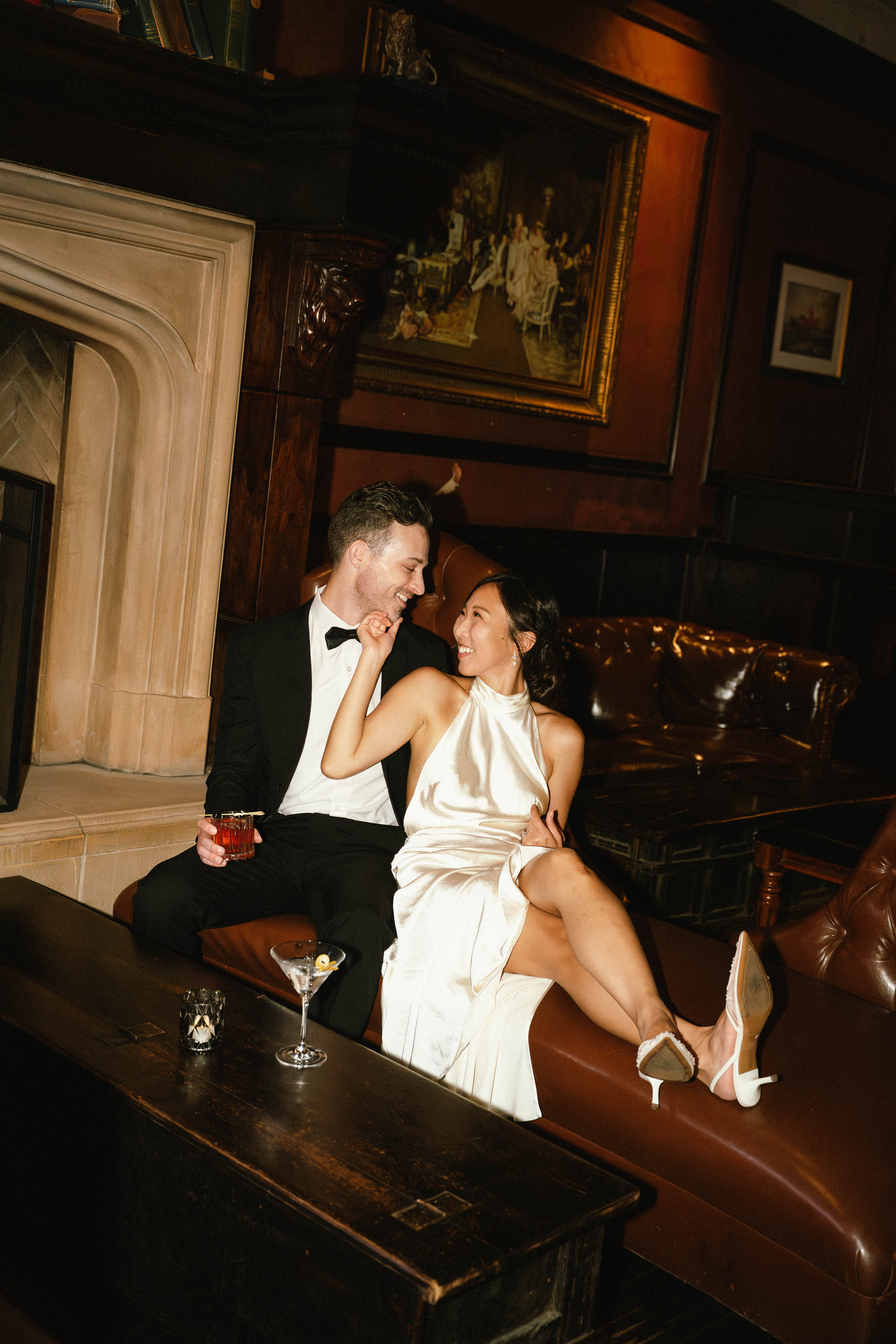 Elegant couple in formal attire relaxing on leather sofa in upscale lounge, woman in white satin gown, man in tuxedo.