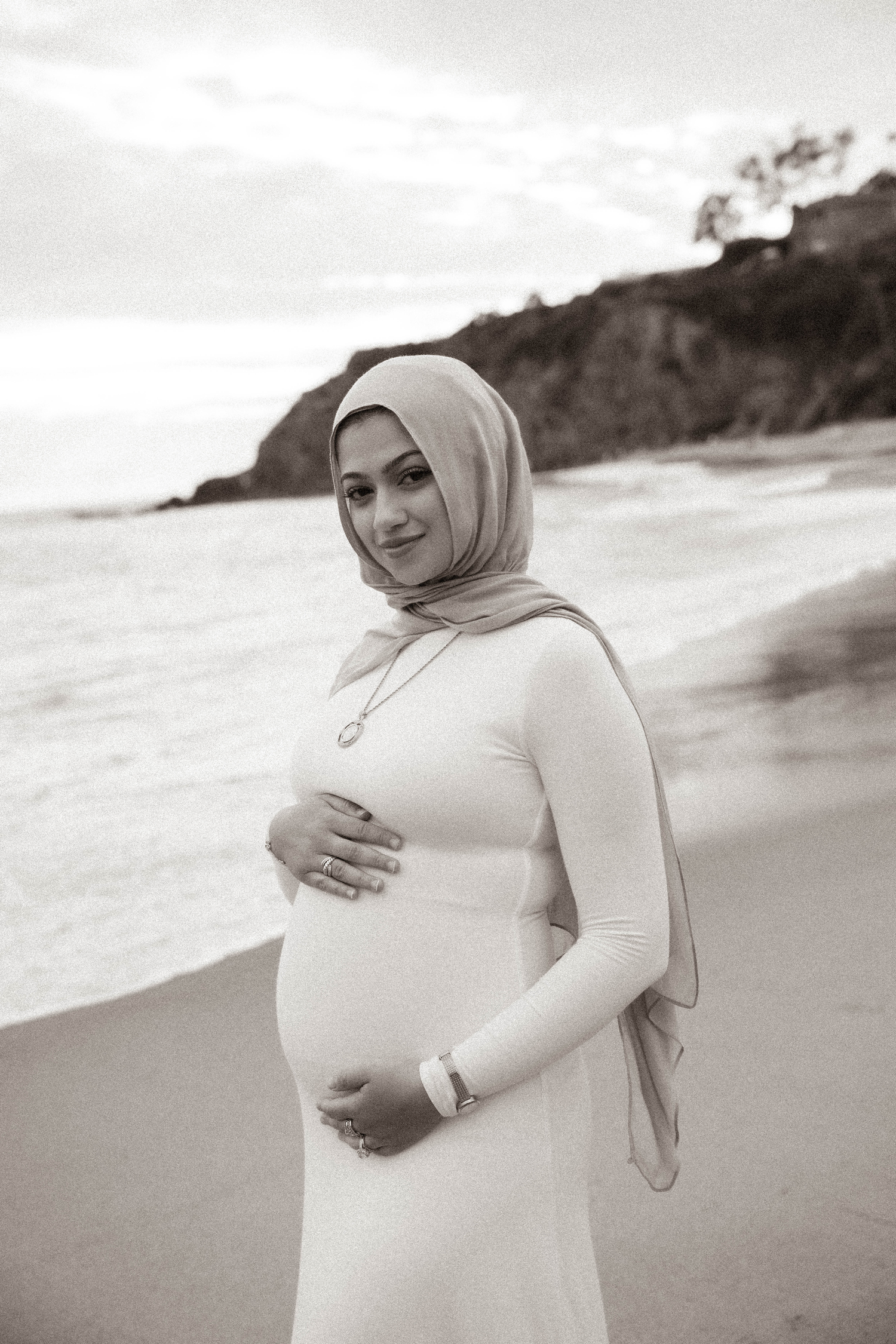 Pregnant woman in hijab and white dress cradling baby bump on beach in black and white maternity photo.
