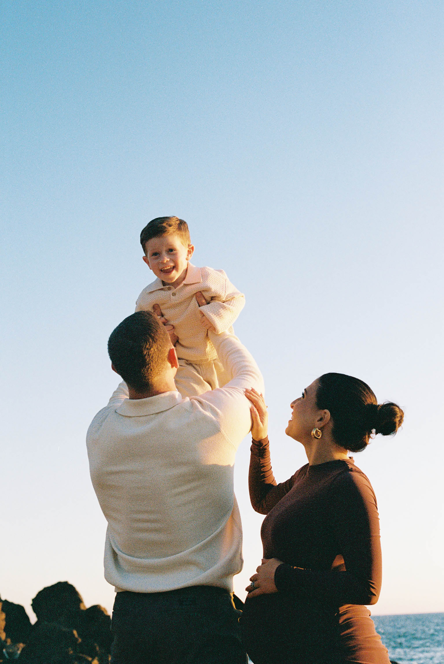 A family lifts their young child up against a clear blue sky near the ocean at golden hour.