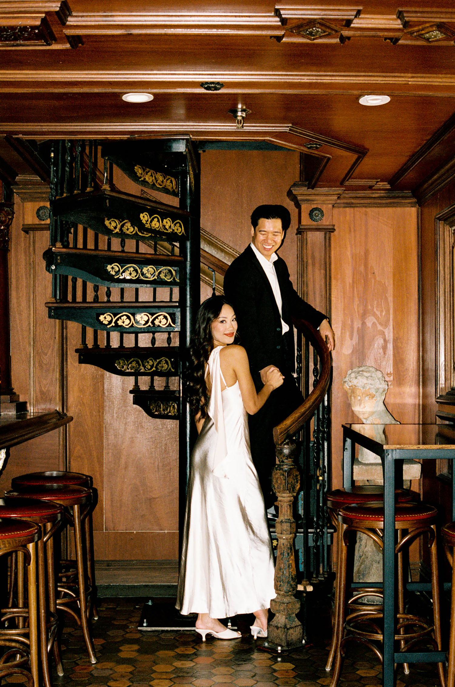 Elegant couple posing by ornate spiral staircase in a classic wood-paneled restaurant interior.