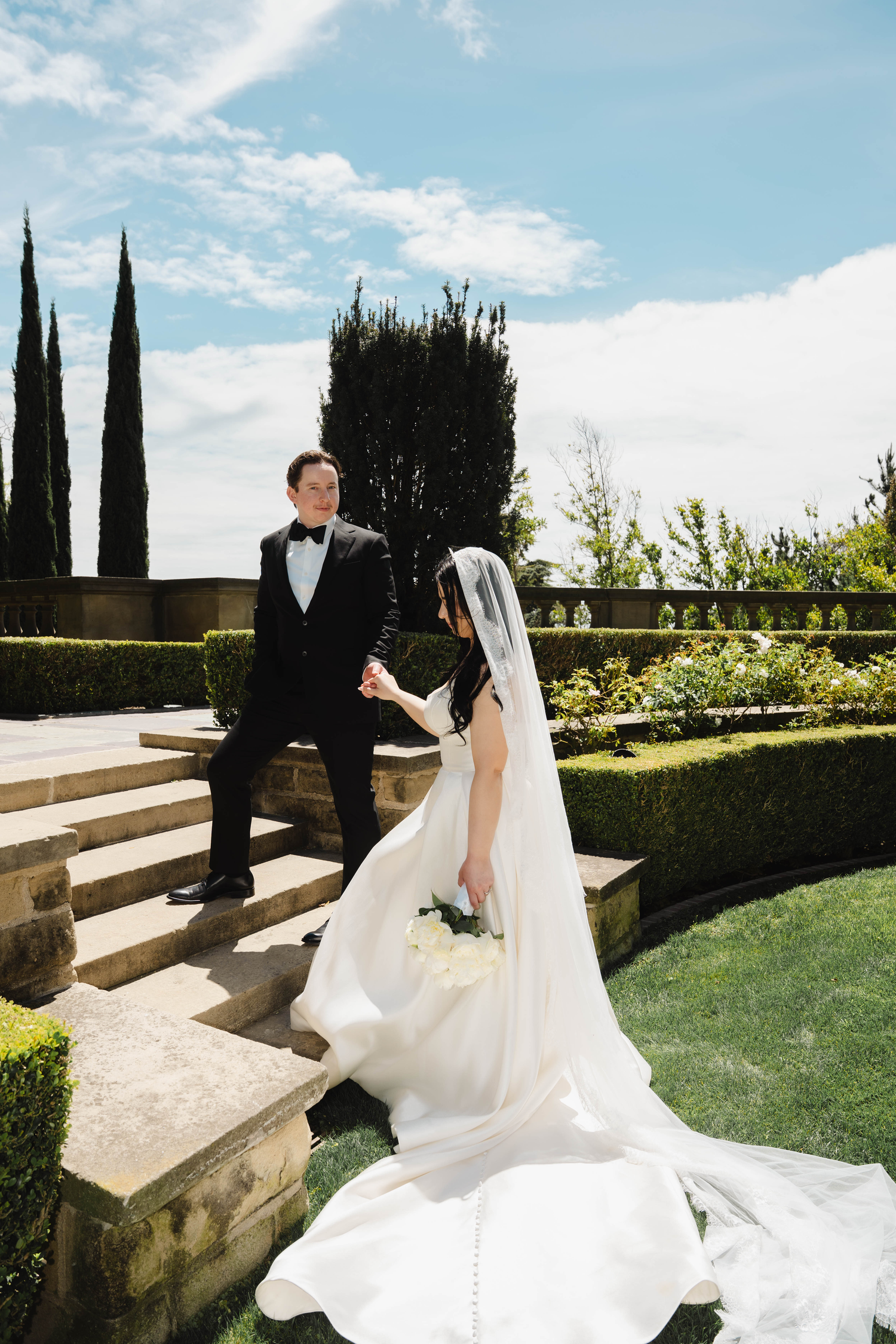 Bride in white gown and veil holds groom's hand on garden steps, surrounded by manicured hedges and cypress trees.