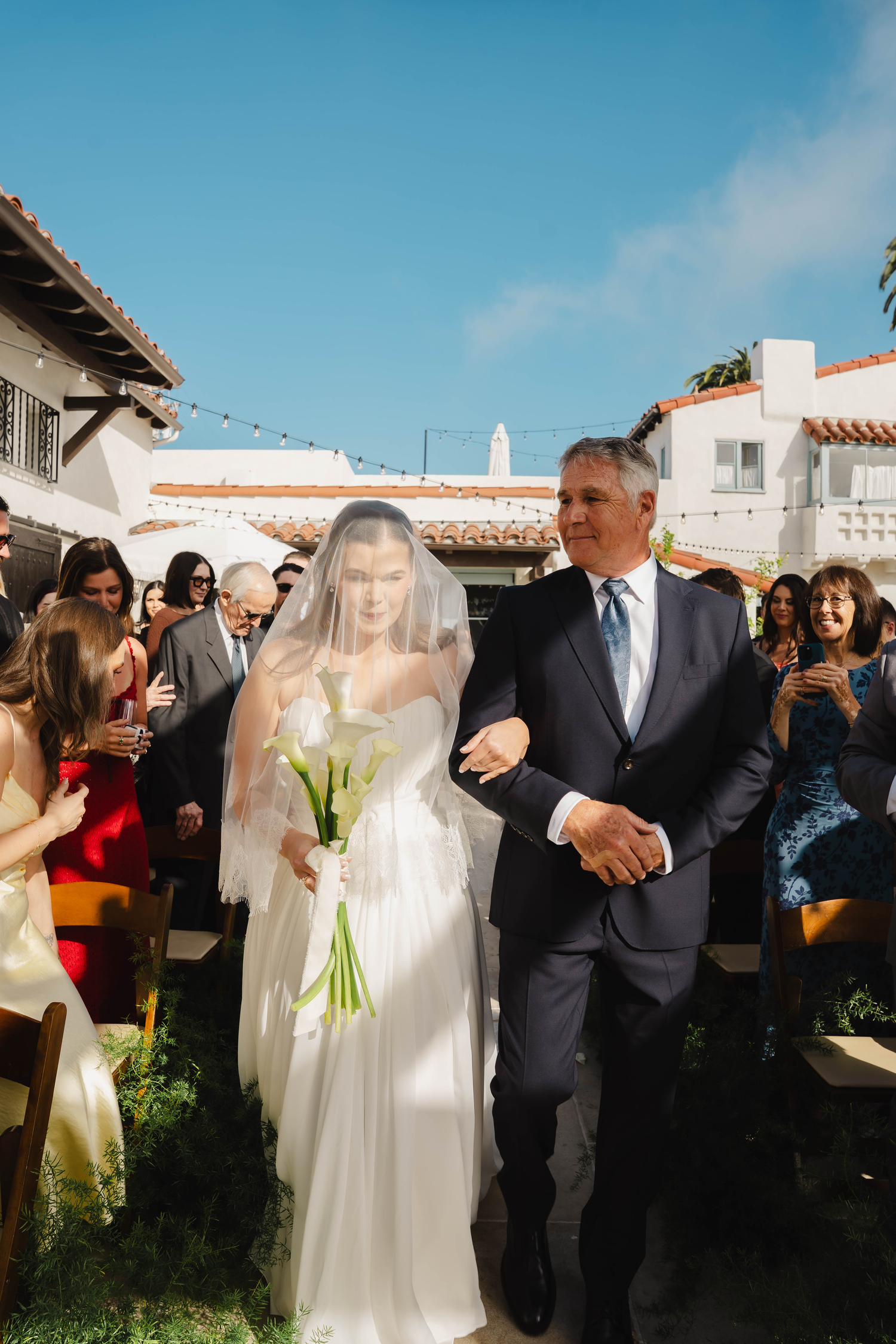 Bride in white gown and veil holding calla lilies walks down outdoor aisle with suited man at sunny wedding.