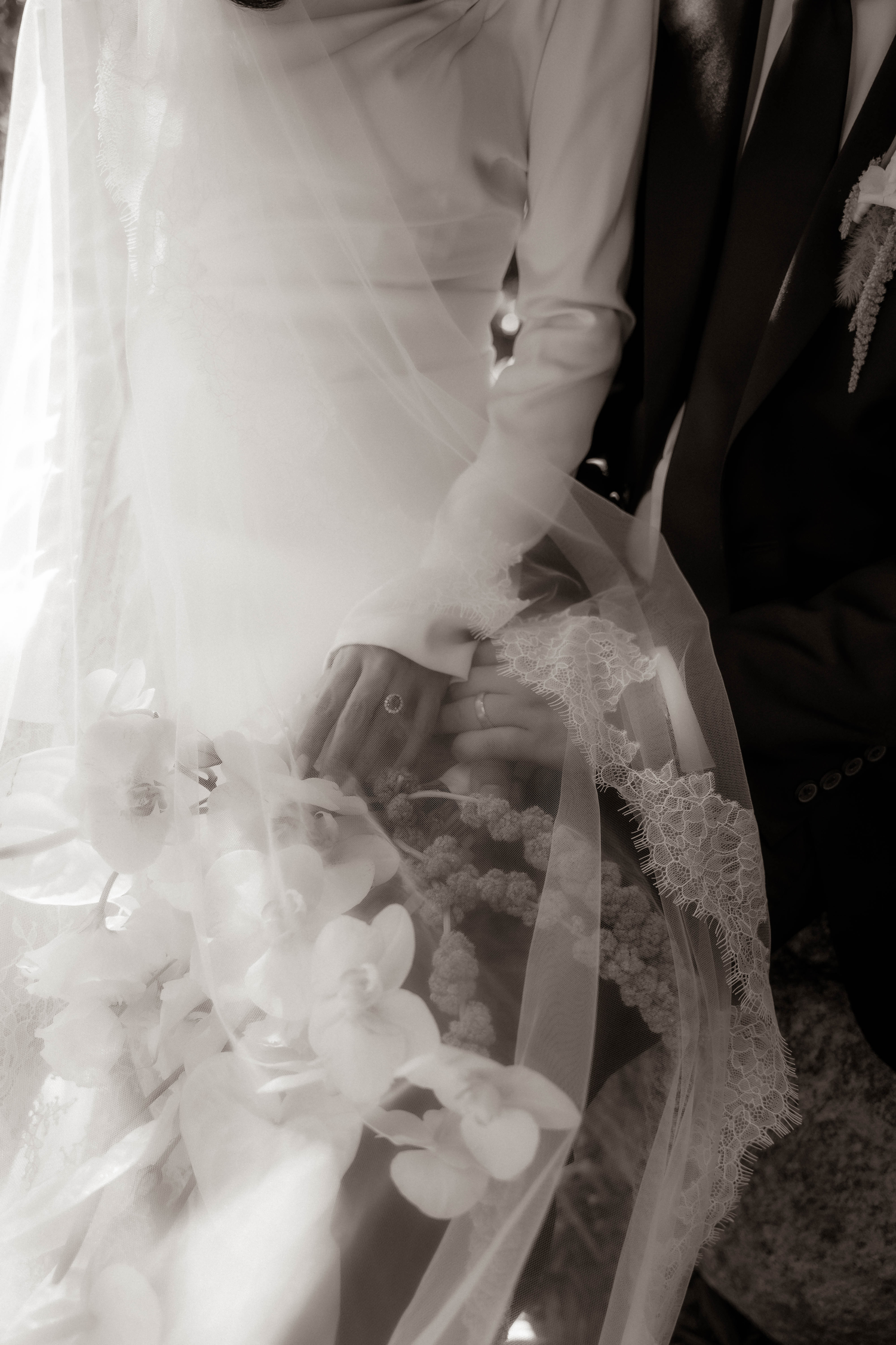 Black and white photo of bride and groom holding hands, bridal bouquet of orchids visible beneath flowing veil.