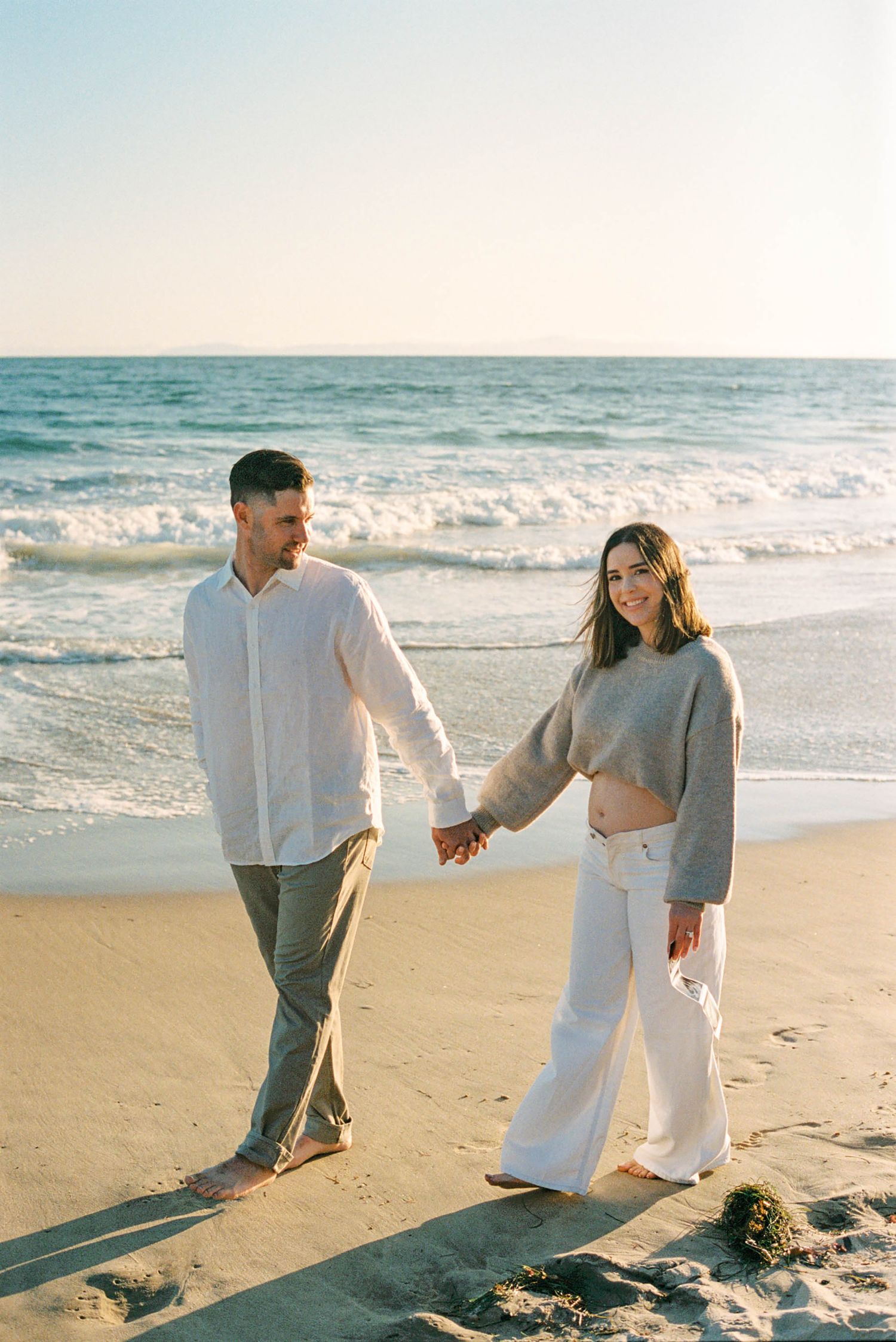 Couple holding hands and walking barefoot on a sandy beach at sunset with ocean waves in the background.