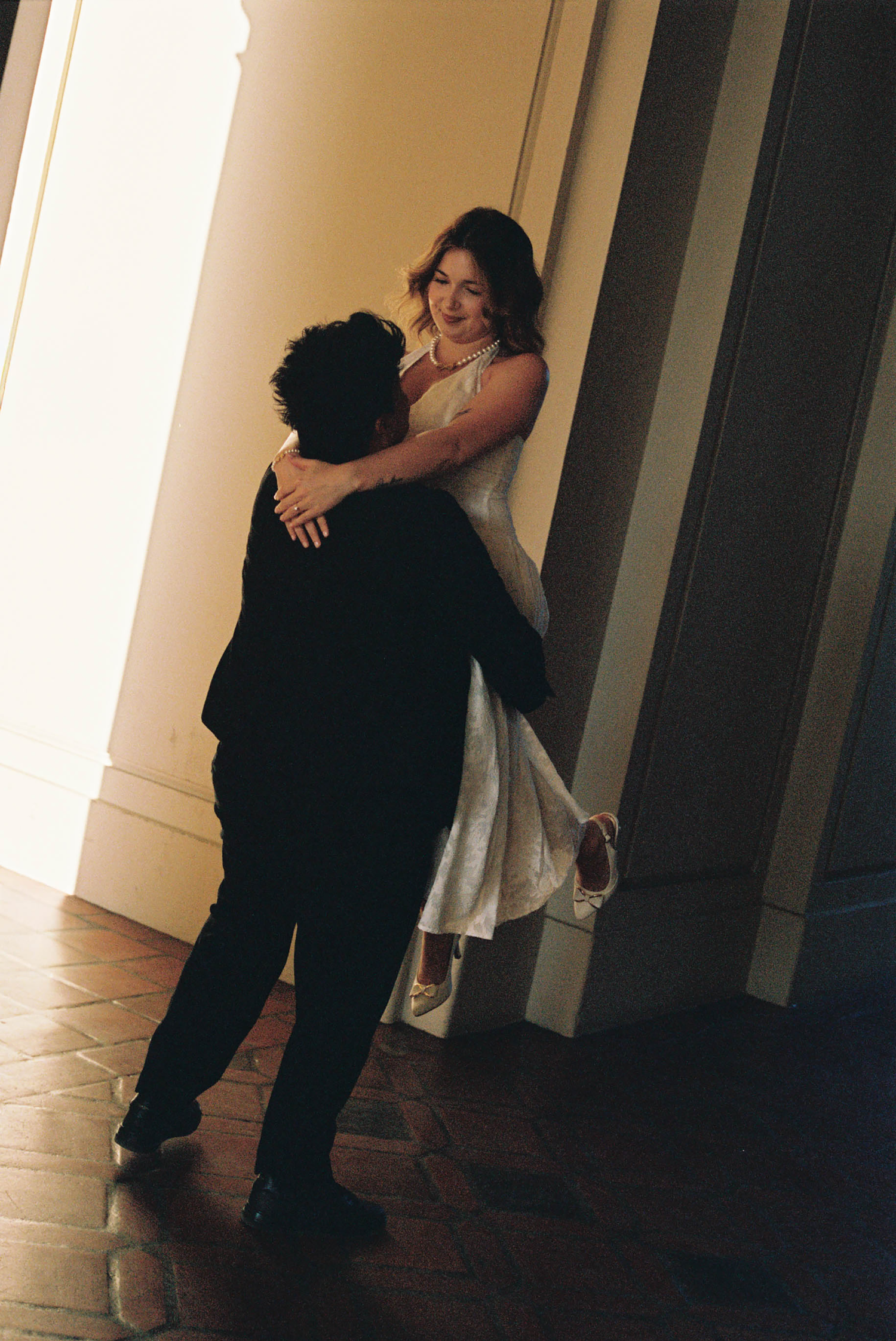 A couple shares a romantic dance embrace in a sunlit hallway, bride in white dress and groom in black suit.