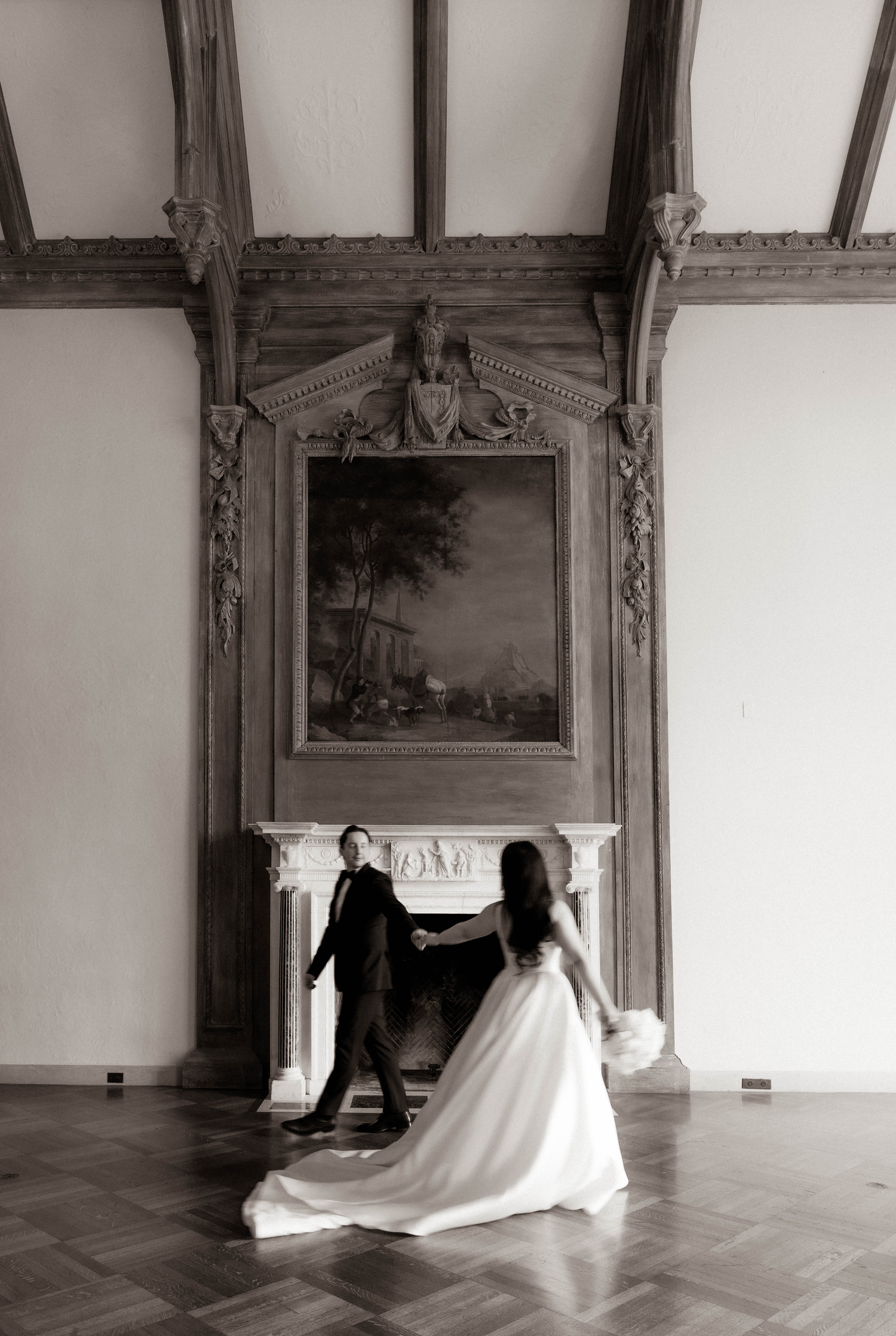 Black and white photo of bride and groom holding hands near ornate fireplace in elegant historic venue.
