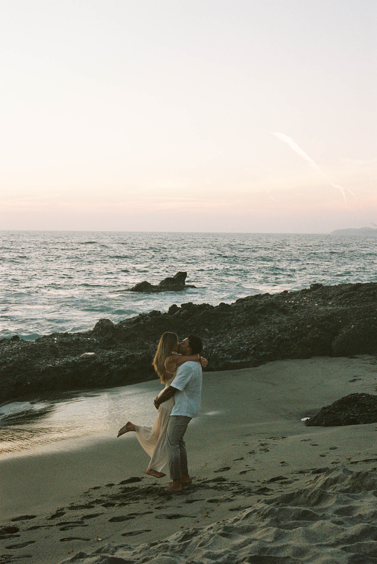 Couple kissing on rocky beach at sunset with ocean waves behind them.