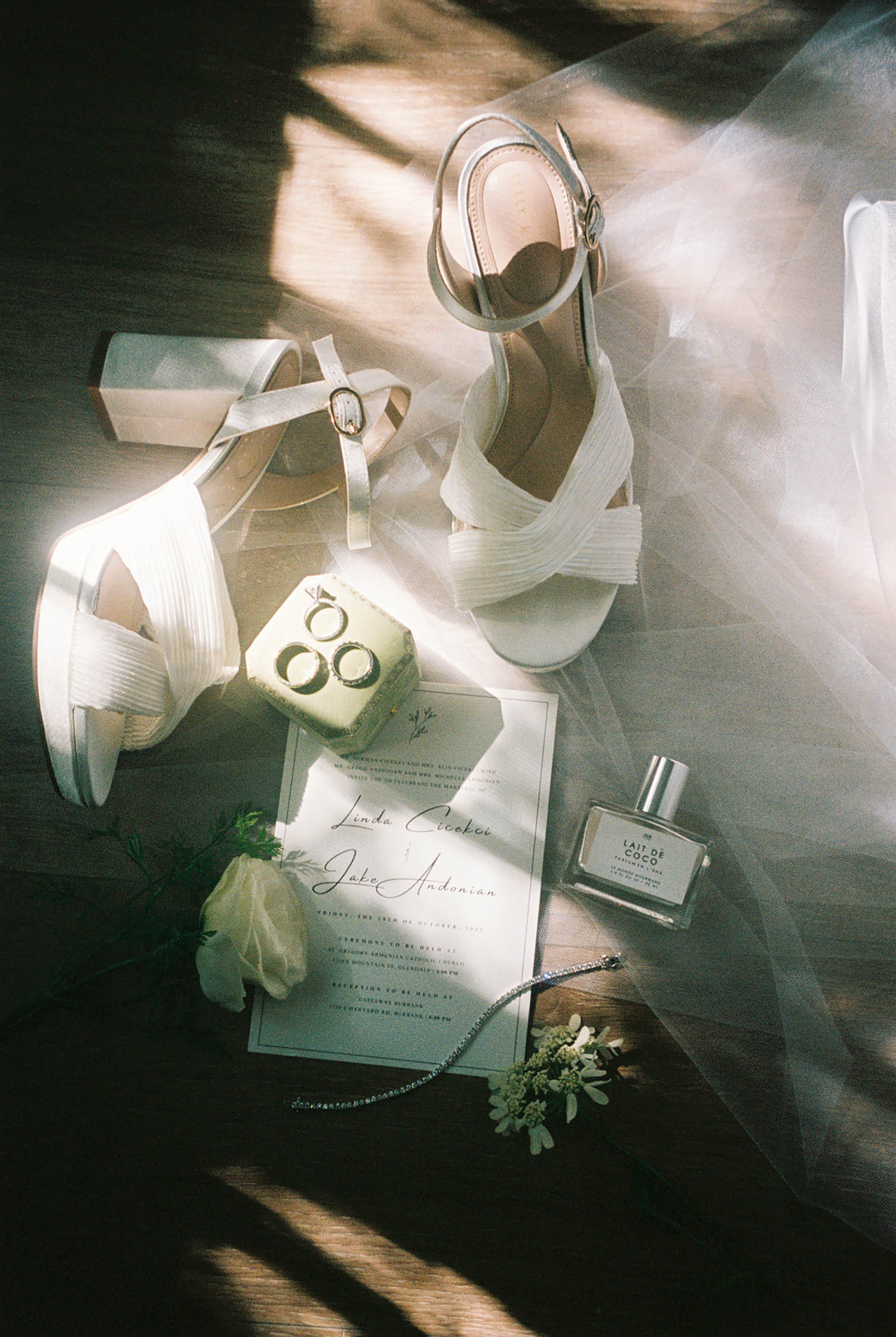 Wedding flat lay with white heels, gold rings, invitation, boutonniere, and perfume in natural light.
