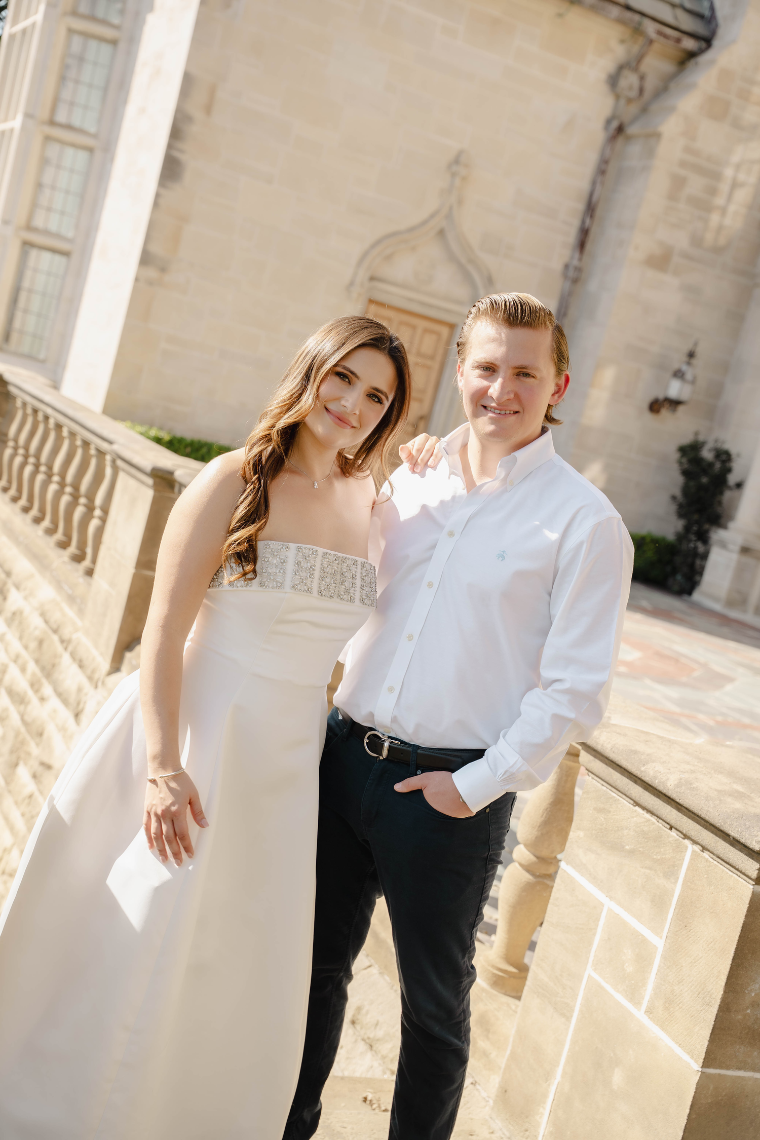 Smiling bride and groom pose together on stone terrace outside a classic architectural building. Greystone Mansion & Gardens