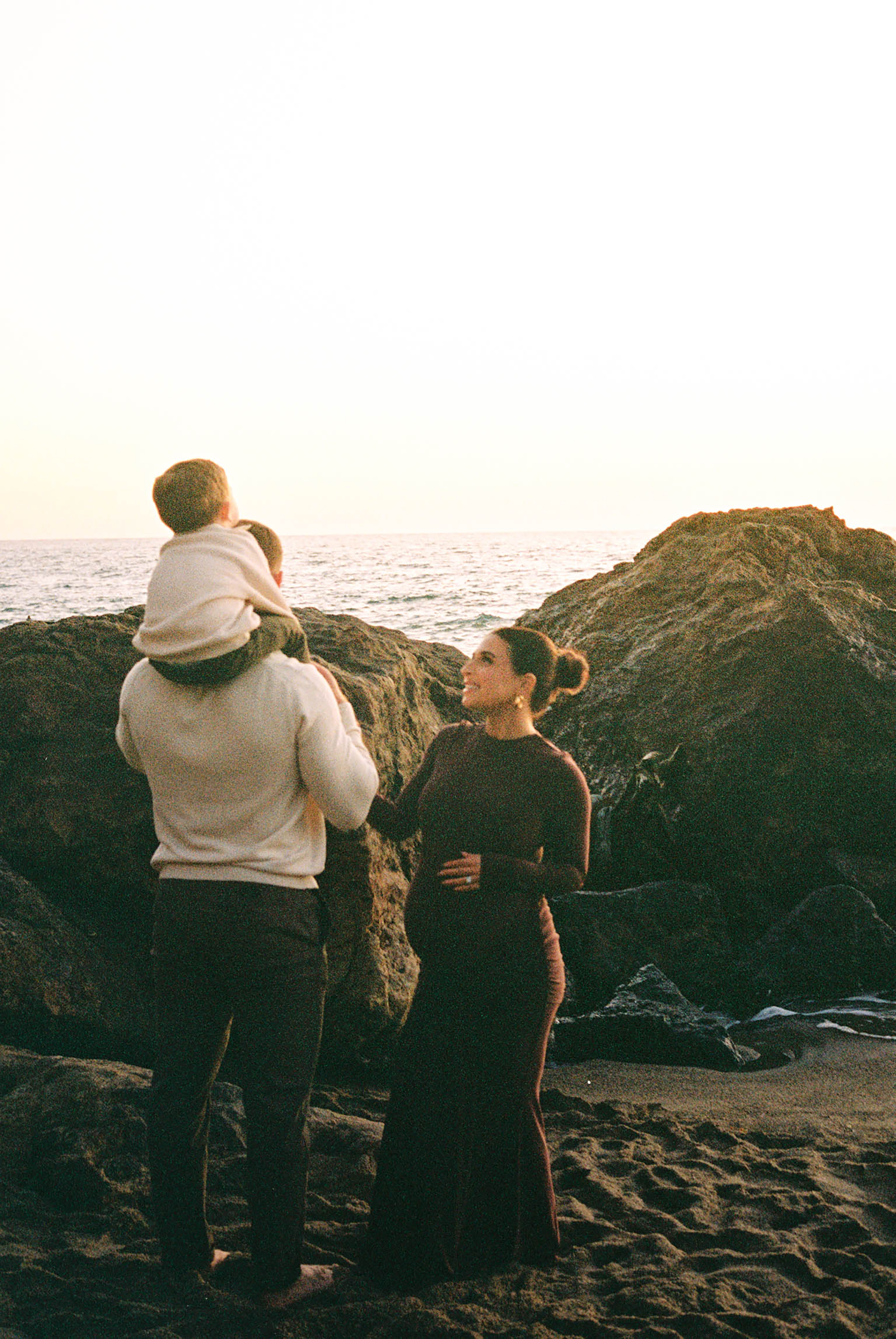 Family of three poses on rocky beach at sunset, man holds child on shoulders while pregnant woman smiles warmly.
