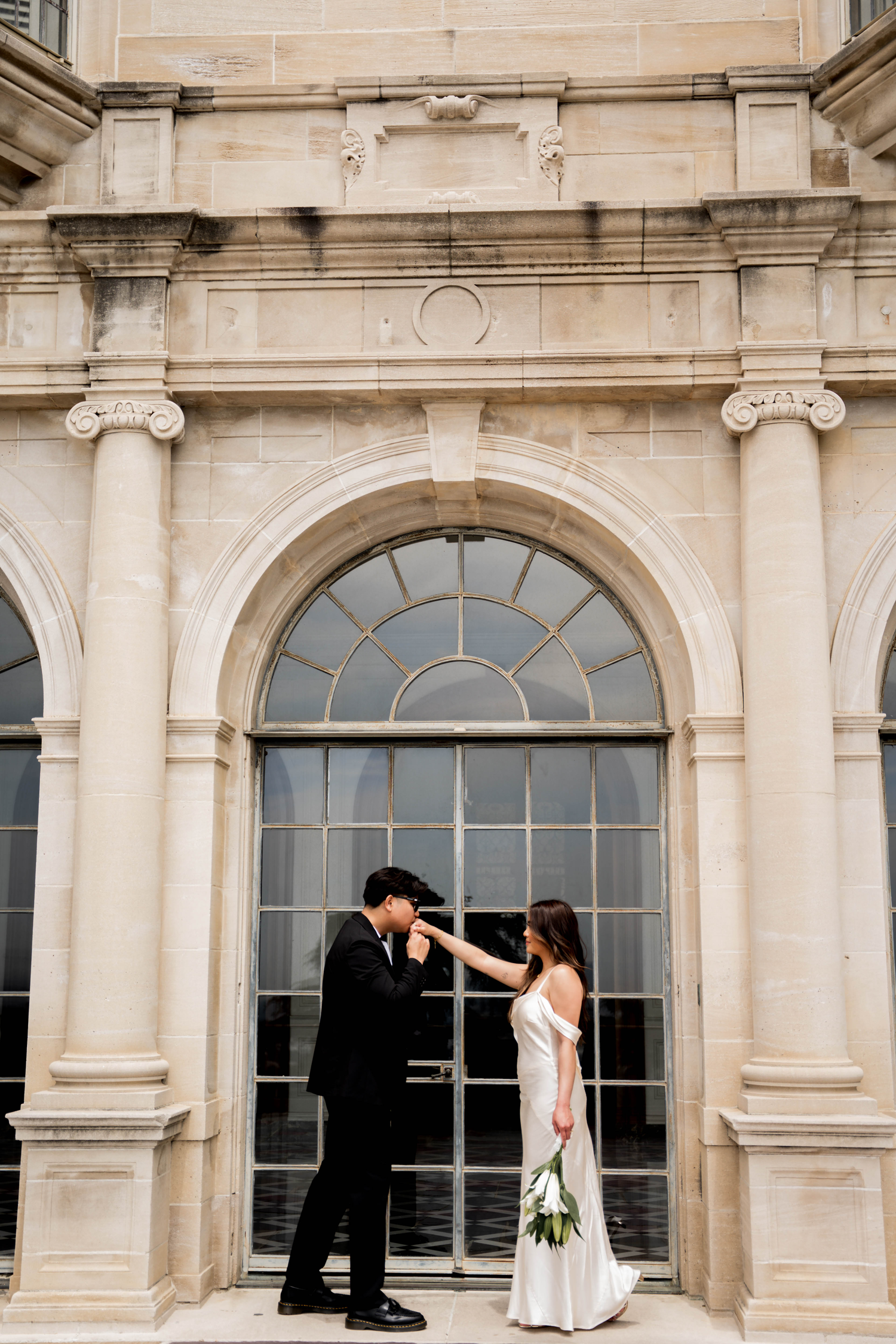 Bride and groom share romantic moment before elegant stone arch building on wedding day.