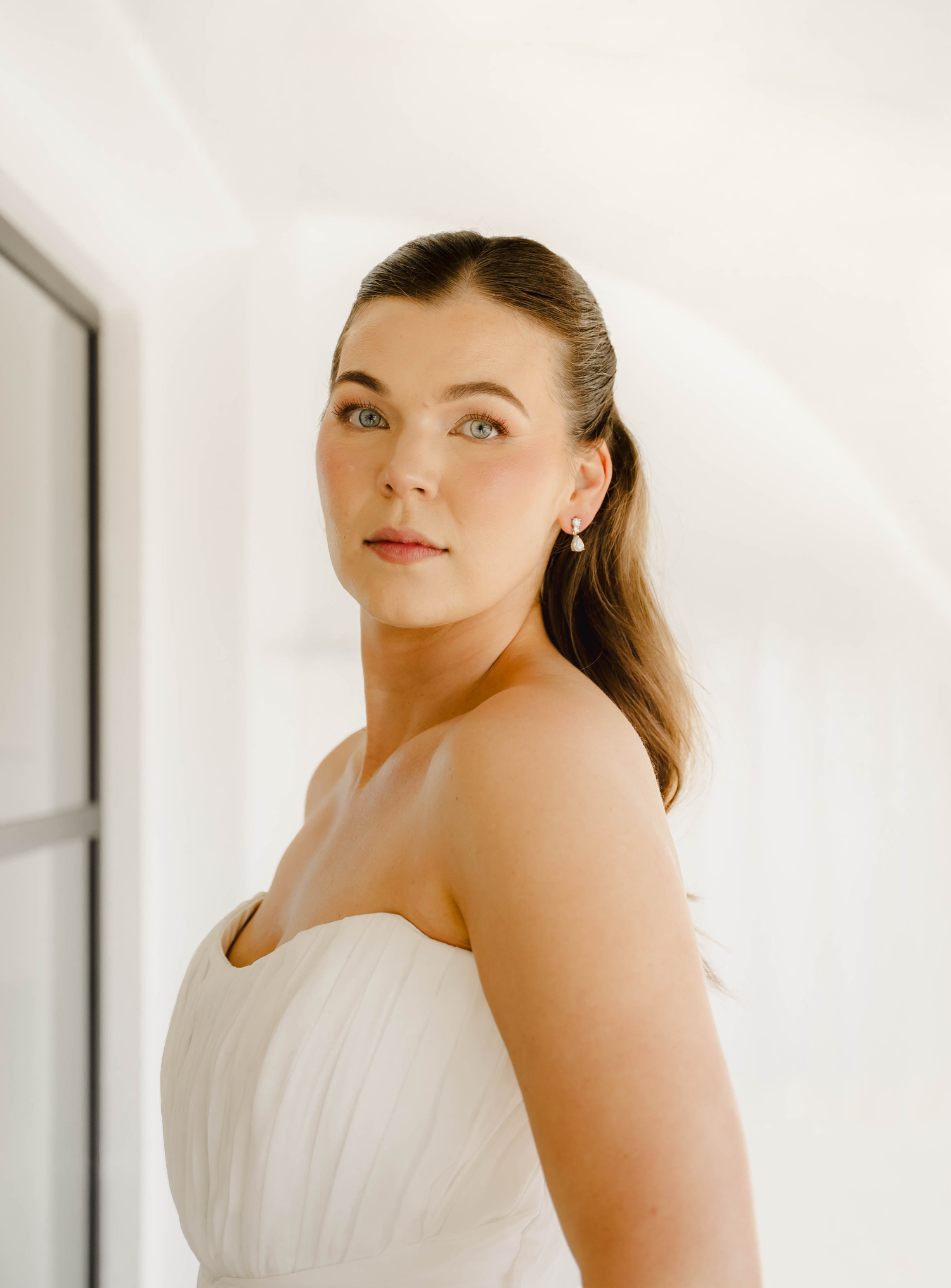 A bride in a strapless white gown with pearl earrings gazes upward in soft natural light.