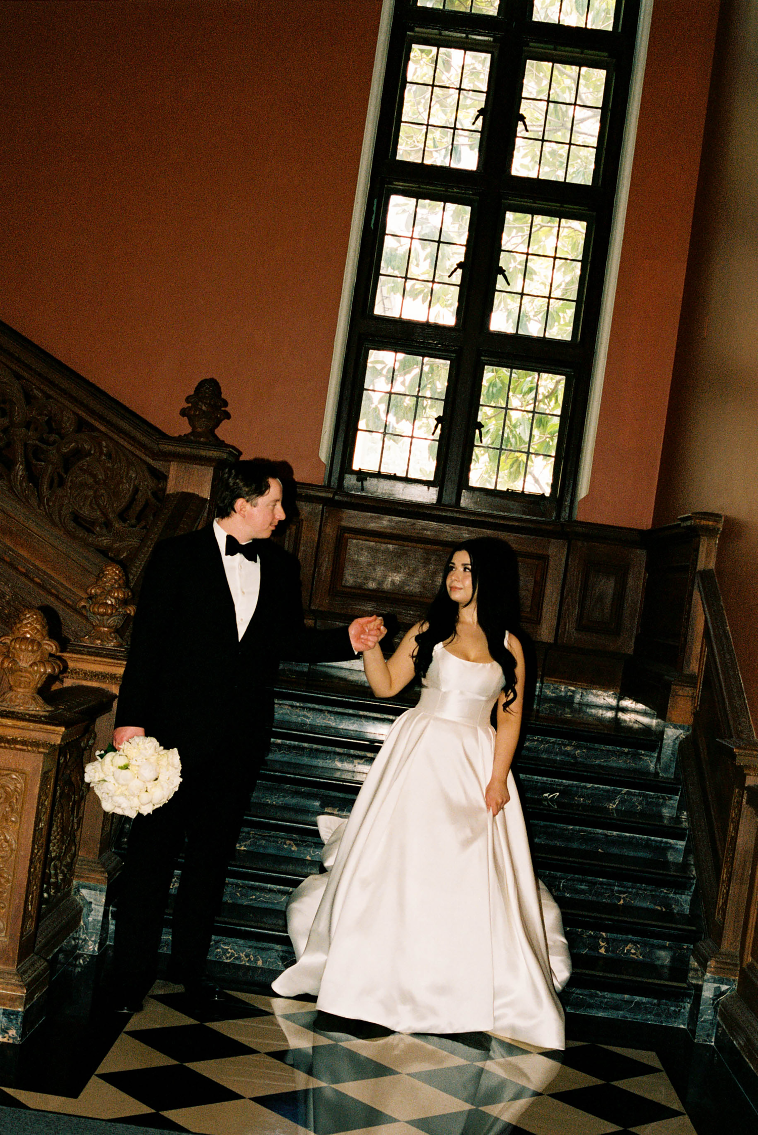 Bride and groom holding hands on grand marble staircase, she in white ball gown, he in black tuxedo, elegant venue.