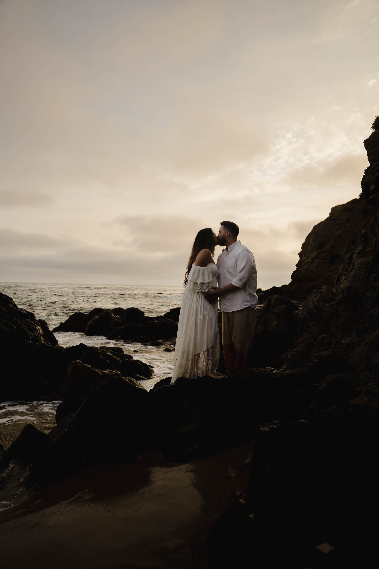 Couple sharing a kiss on rocky coastal cliffs at golden hour, maternity photos.
