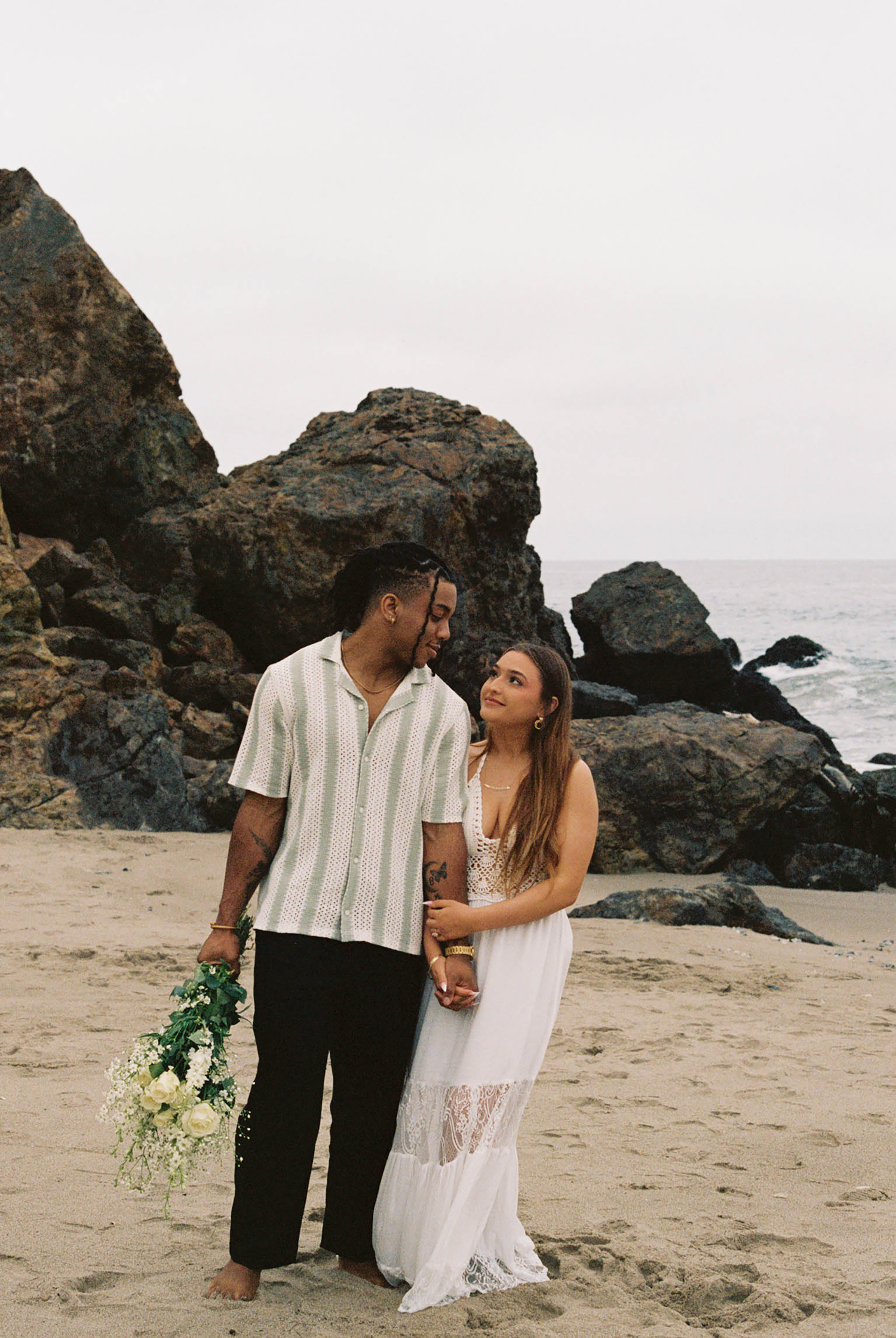 Couple standing on a sandy beach with rocky cliffs, man holding white flowers, woman in white dress, ocean backdrop.