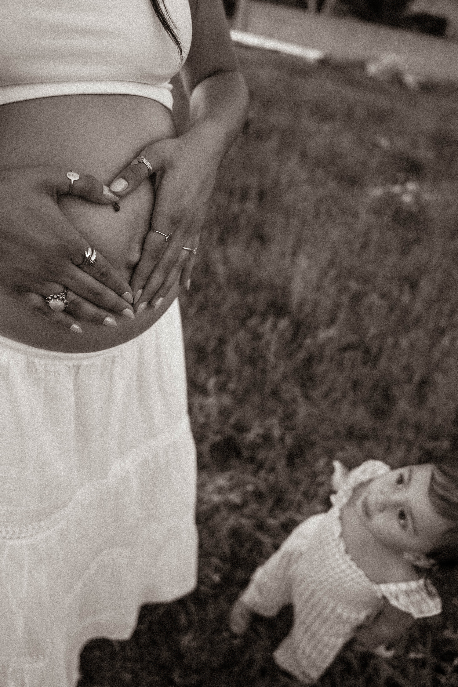 Black and white maternity photo of a pregnant woman cradling her belly while her toddler looks up at her outdoors.