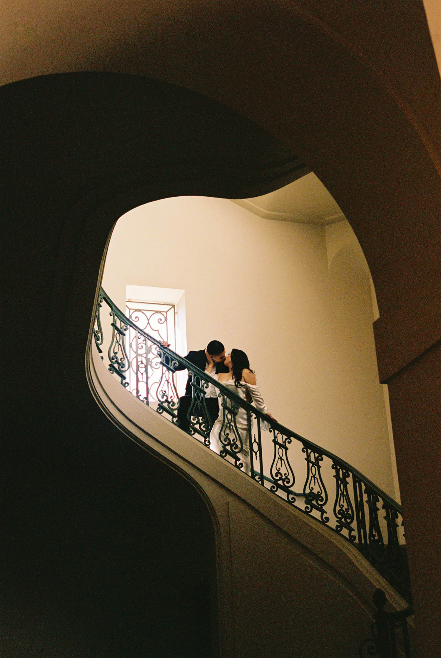 Couple shares romantic kiss on elegant curved staircase with ornate iron railing in grand building. Italy Vibes. Pasadena City Hall.
