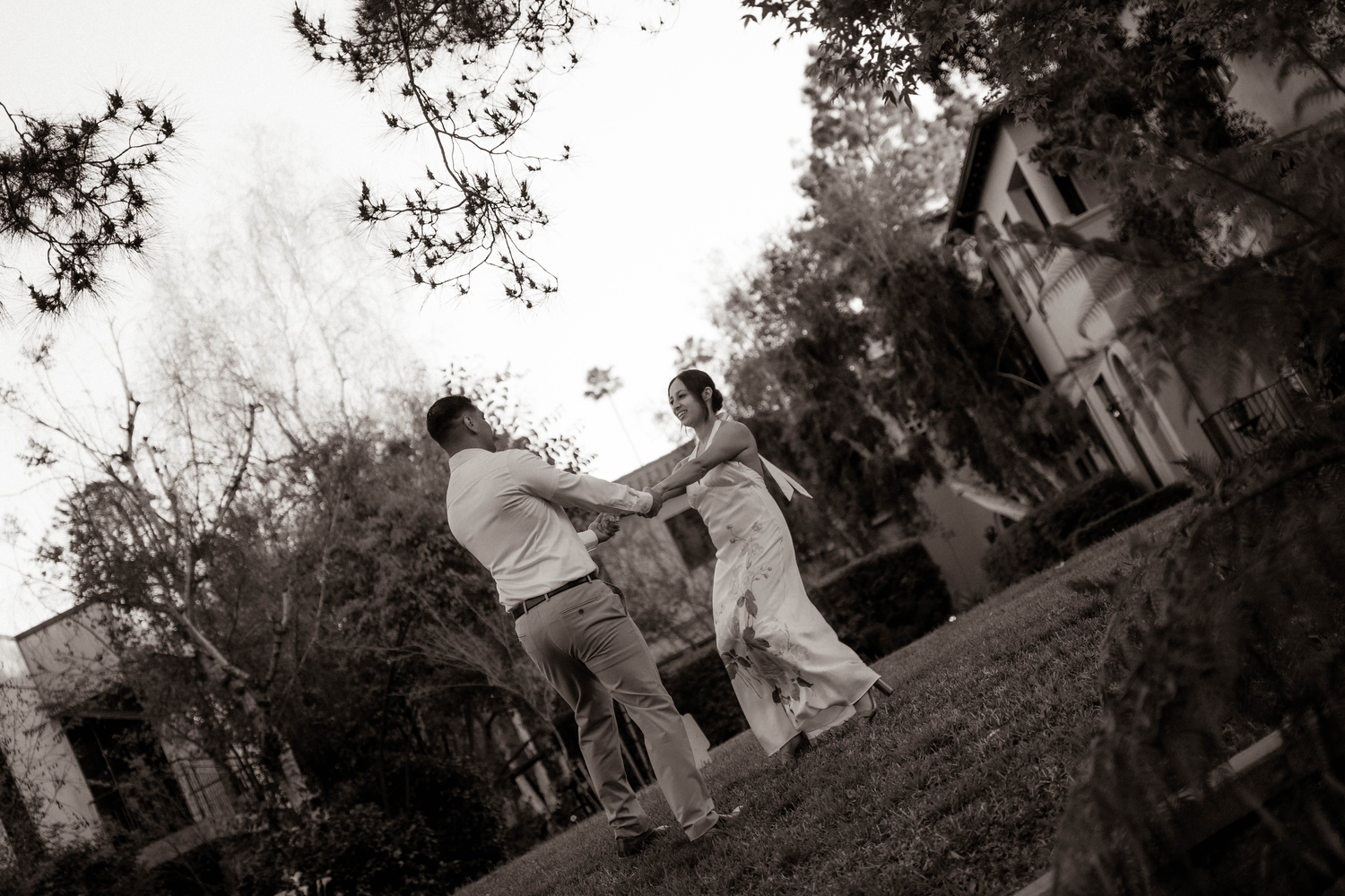 Black and white photo of couple holding hands and dancing outdoors among trees in casual attire.