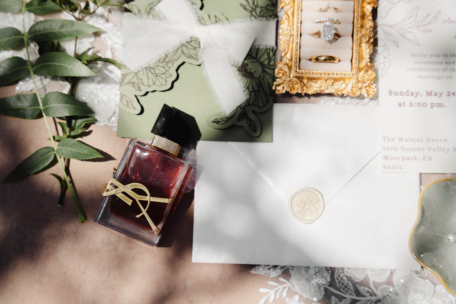 Elegant wedding flat lay with YSL perfume, gold-framed ring, wax seal envelope, green eucalyptus, and sage invitation suite.