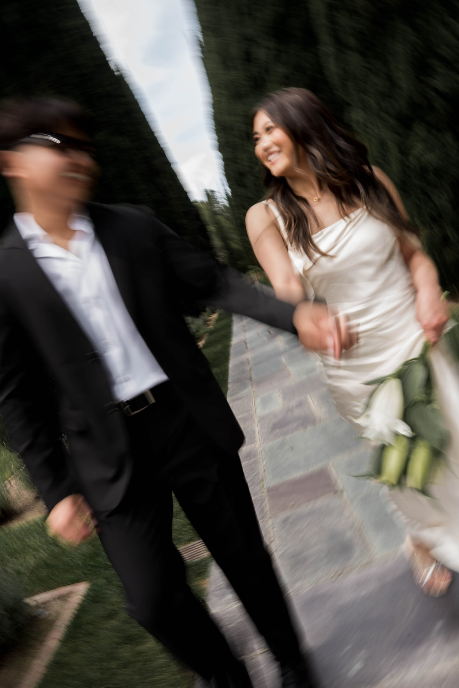 A joyful bride in a white dress holds a bouquet while walking with a groom in a black suit on a garden path.
