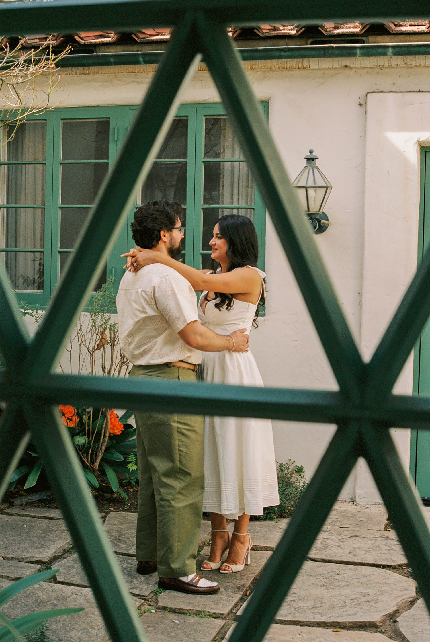 A smiling couple embraces in a courtyard framed by green geometric lattice and French doors.