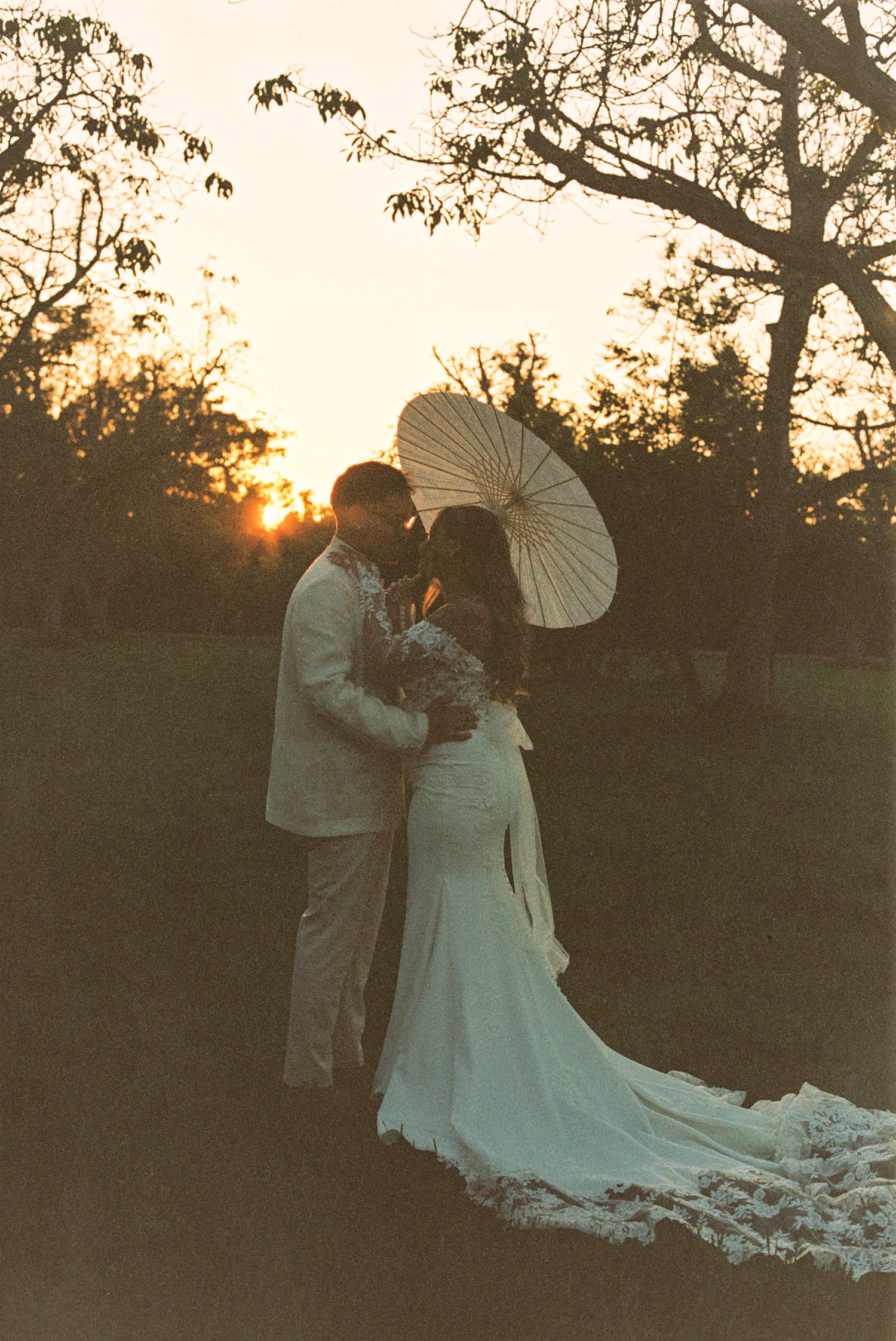 Couple sharing a romantic kiss at sunset, bride in lace gown holding parasol, groom in white suit, golden hour backdrop.