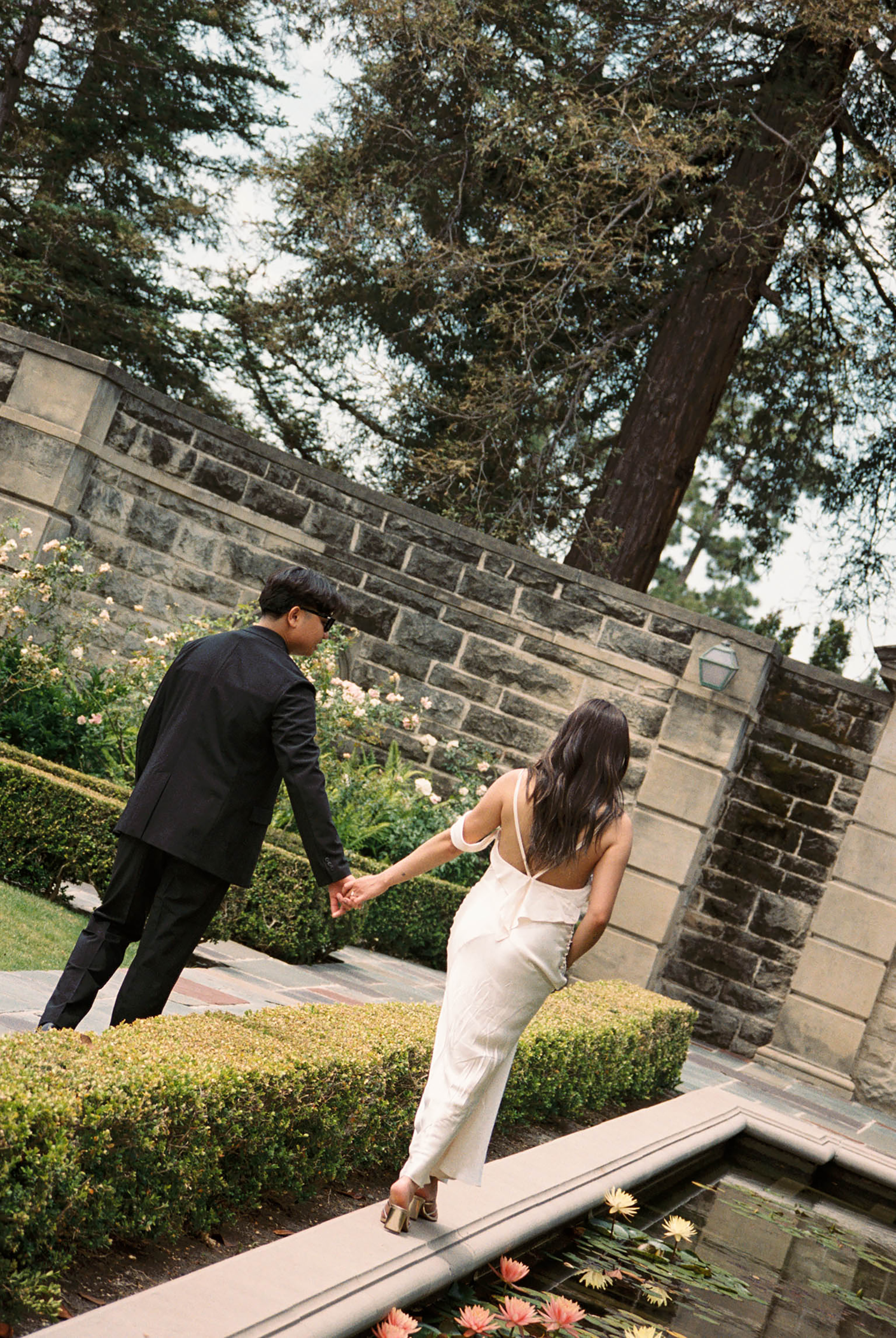 Bride and groom walk hand in hand through a lush garden with stone walls and a reflecting pool.