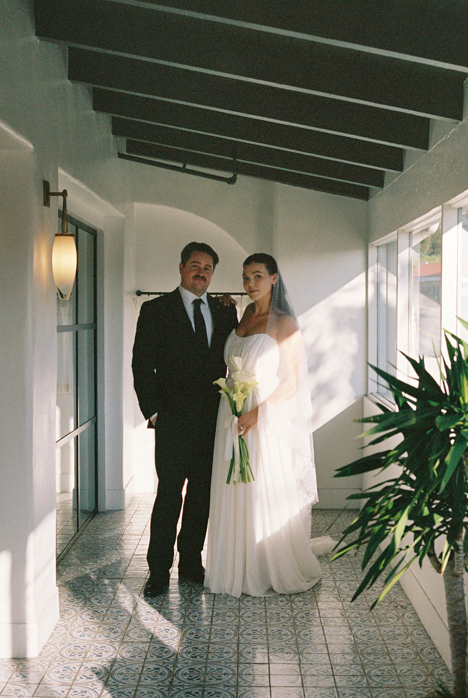 Bride in white gown holding calla lilies stands with groom in black suit in sunlit corridor with tiled floor.