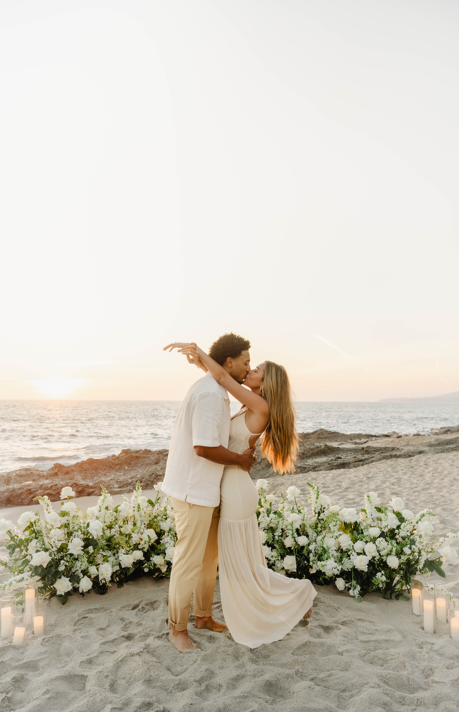 Couple kissing at sunset beach proposal with white floral arrangements and candles on sand by the ocean.