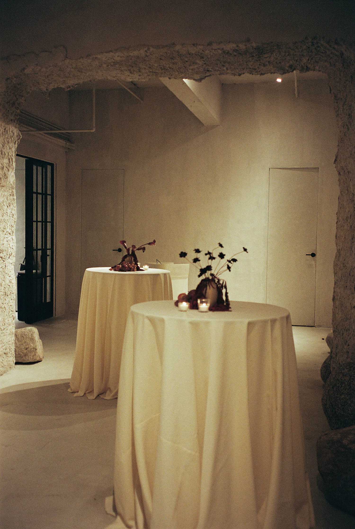 Elegant event space with draped cream cocktail tables, floral arrangements, and warm candlelight in a rustic stone venue.