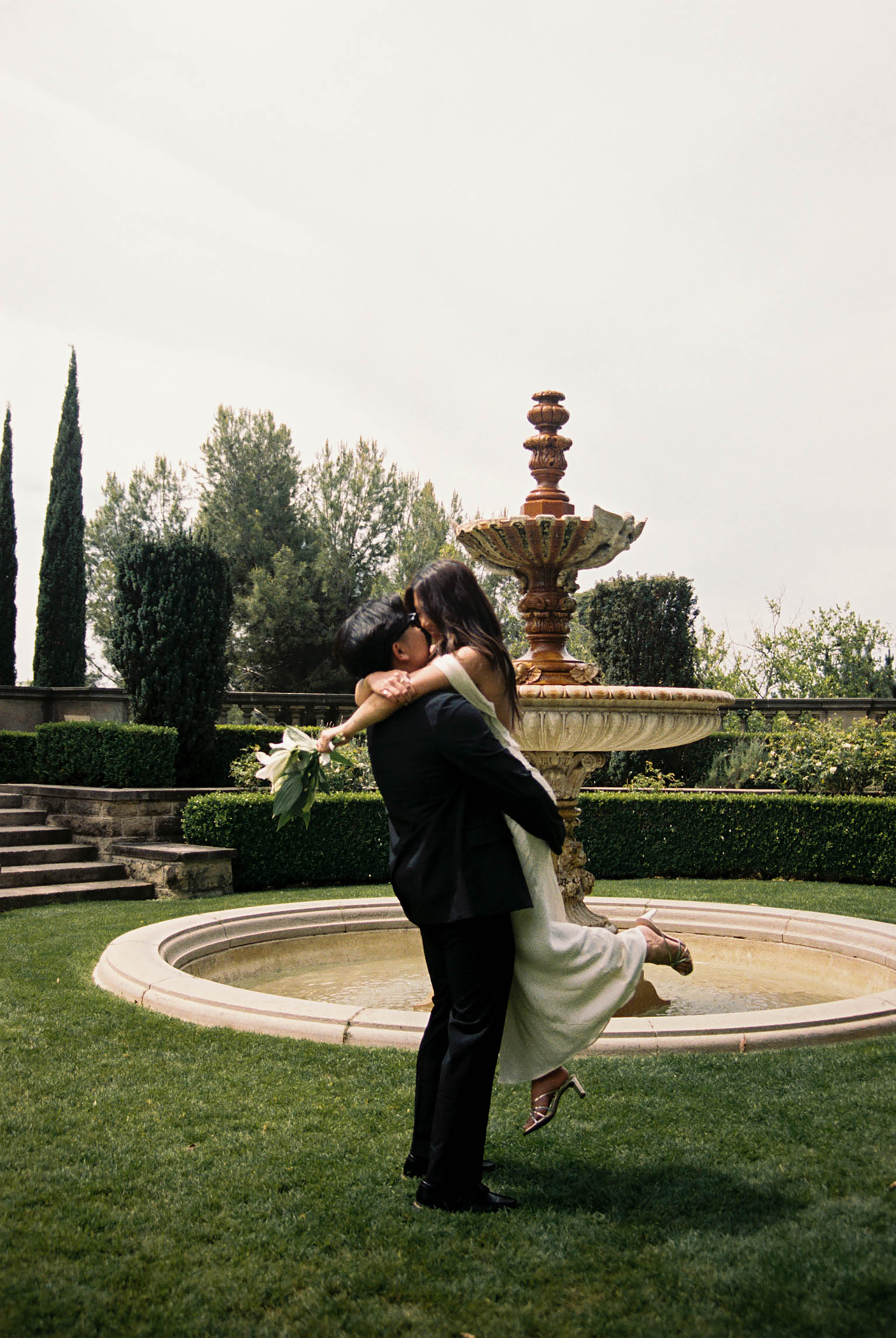 Groom lifting bride in front of ornate stone fountain in lush garden during romantic outdoor wedding celebration.
