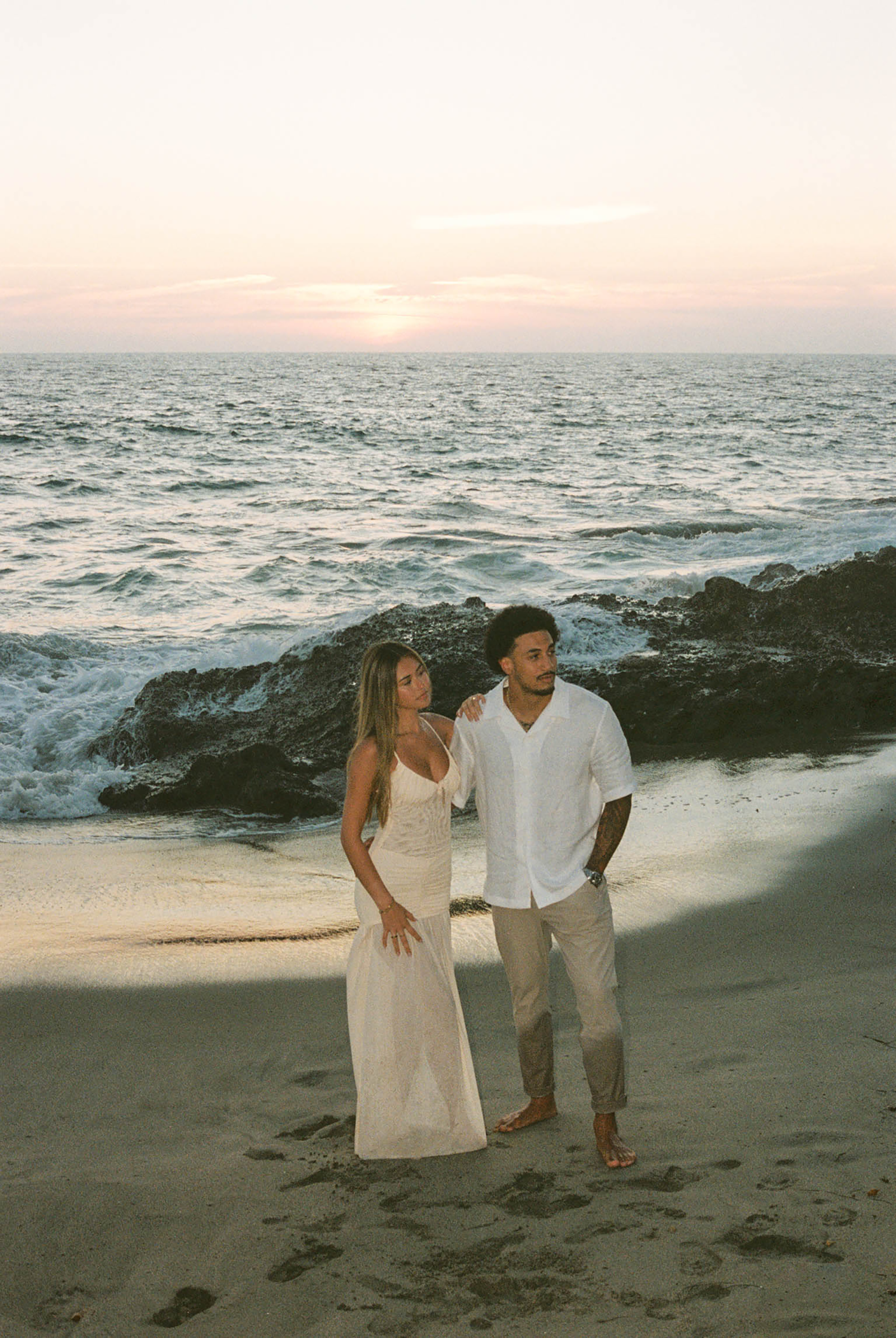 Couple standing together on sandy beach at sunset with ocean waves and rocks behind them.