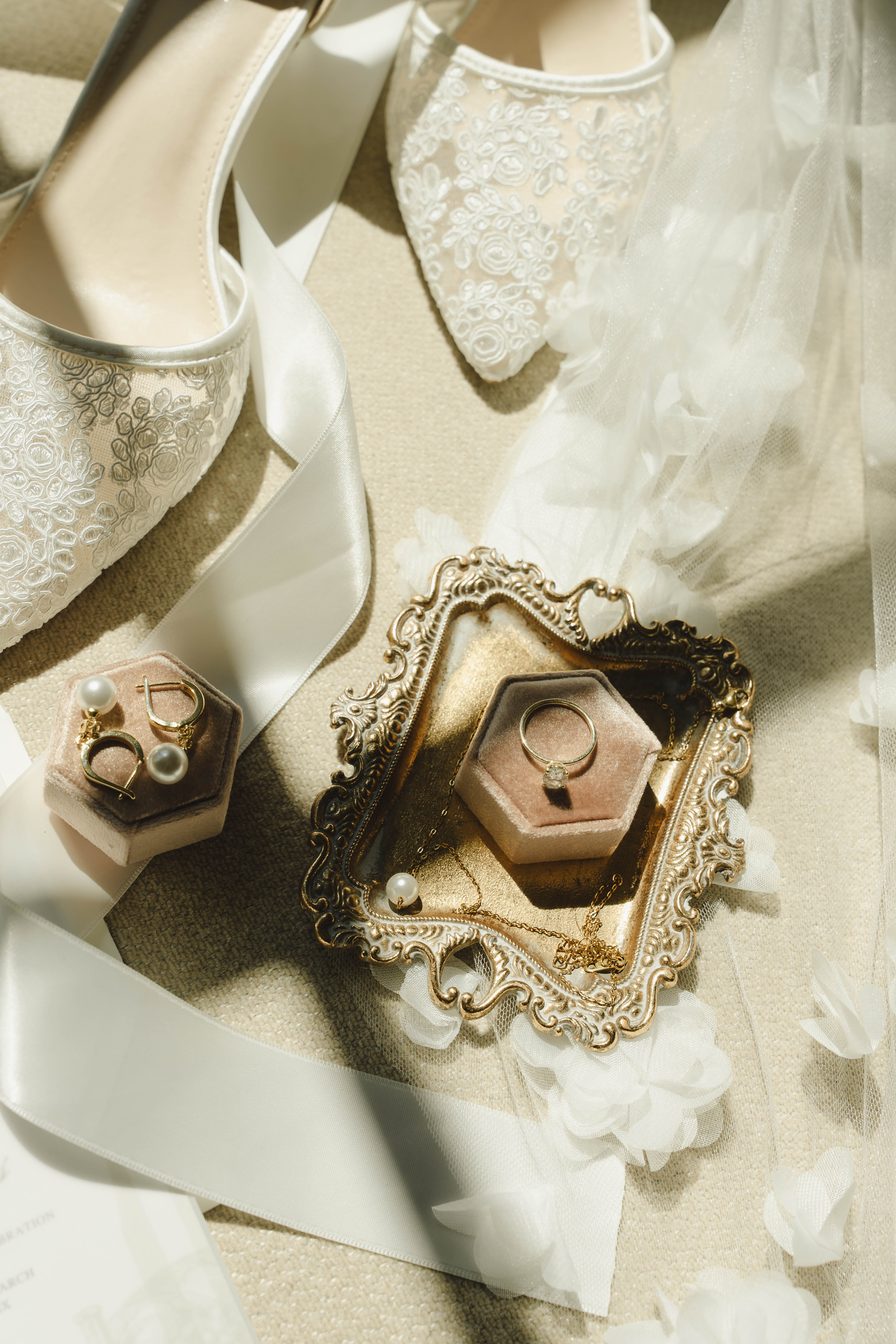 Elegant bridal flat lay featuring lace heels, white ribbon, pearl earrings, gold ring box, and vintage ornate tray.