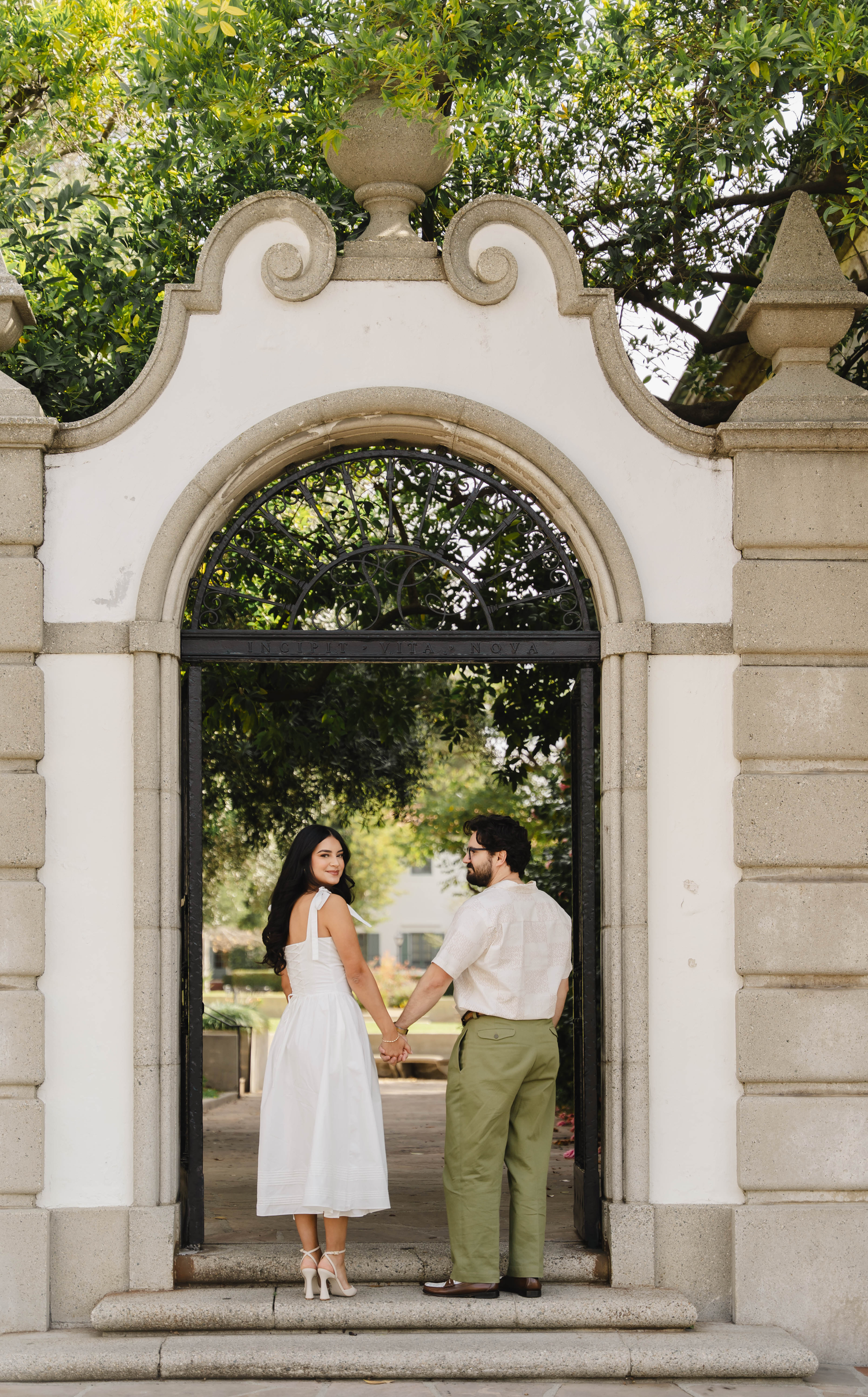 A couple holds hands facing each other beneath an elegant stone archway surrounded by greenery.