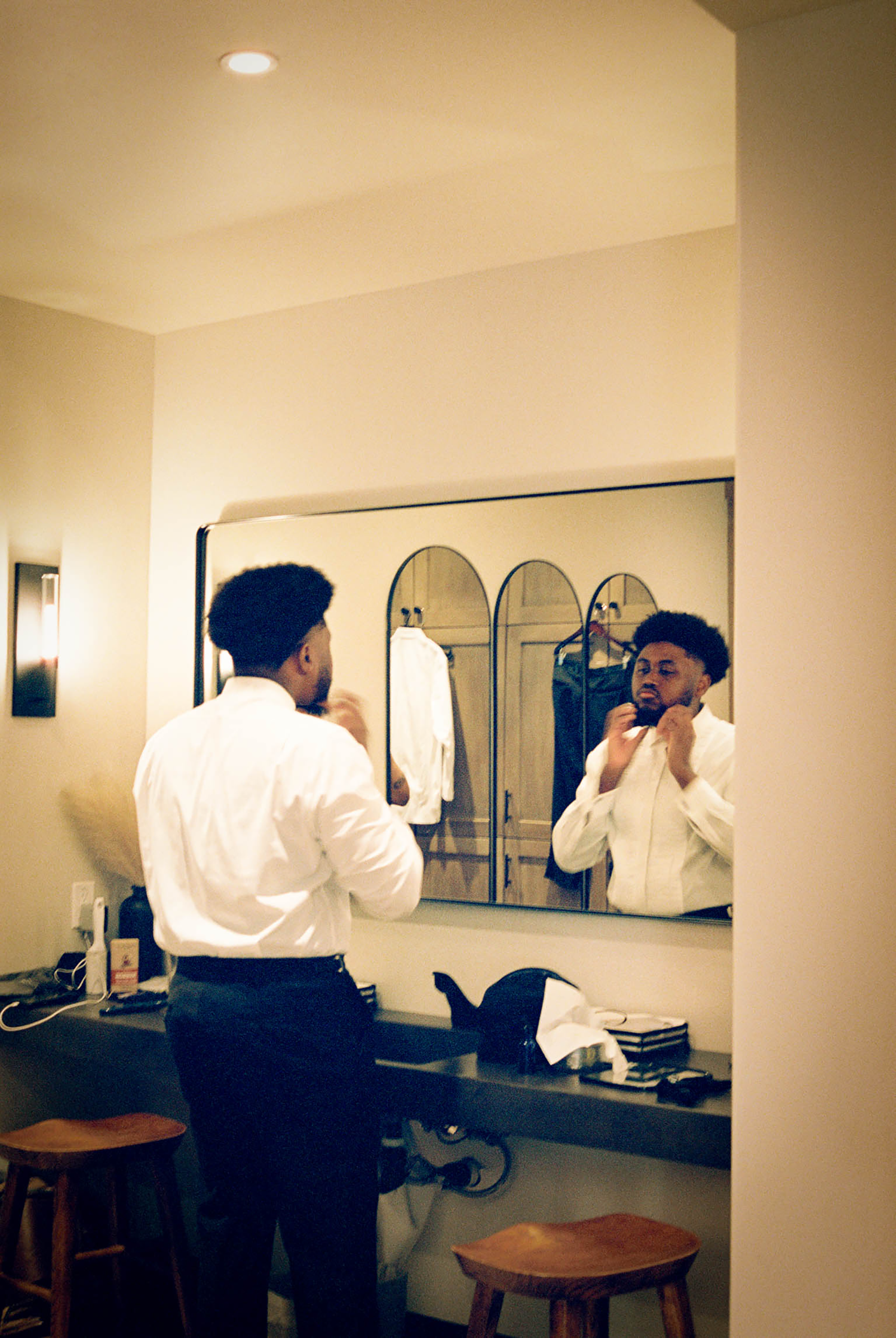 A man in a white dress shirt adjusts his collar while looking in an arched bathroom mirror.