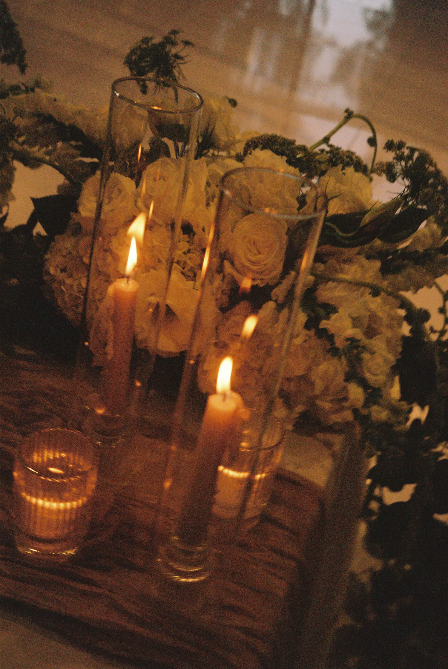 Romantic wedding centerpiece with white roses, greenery, and glowing taper candles in glass holders on a wooden tray.
