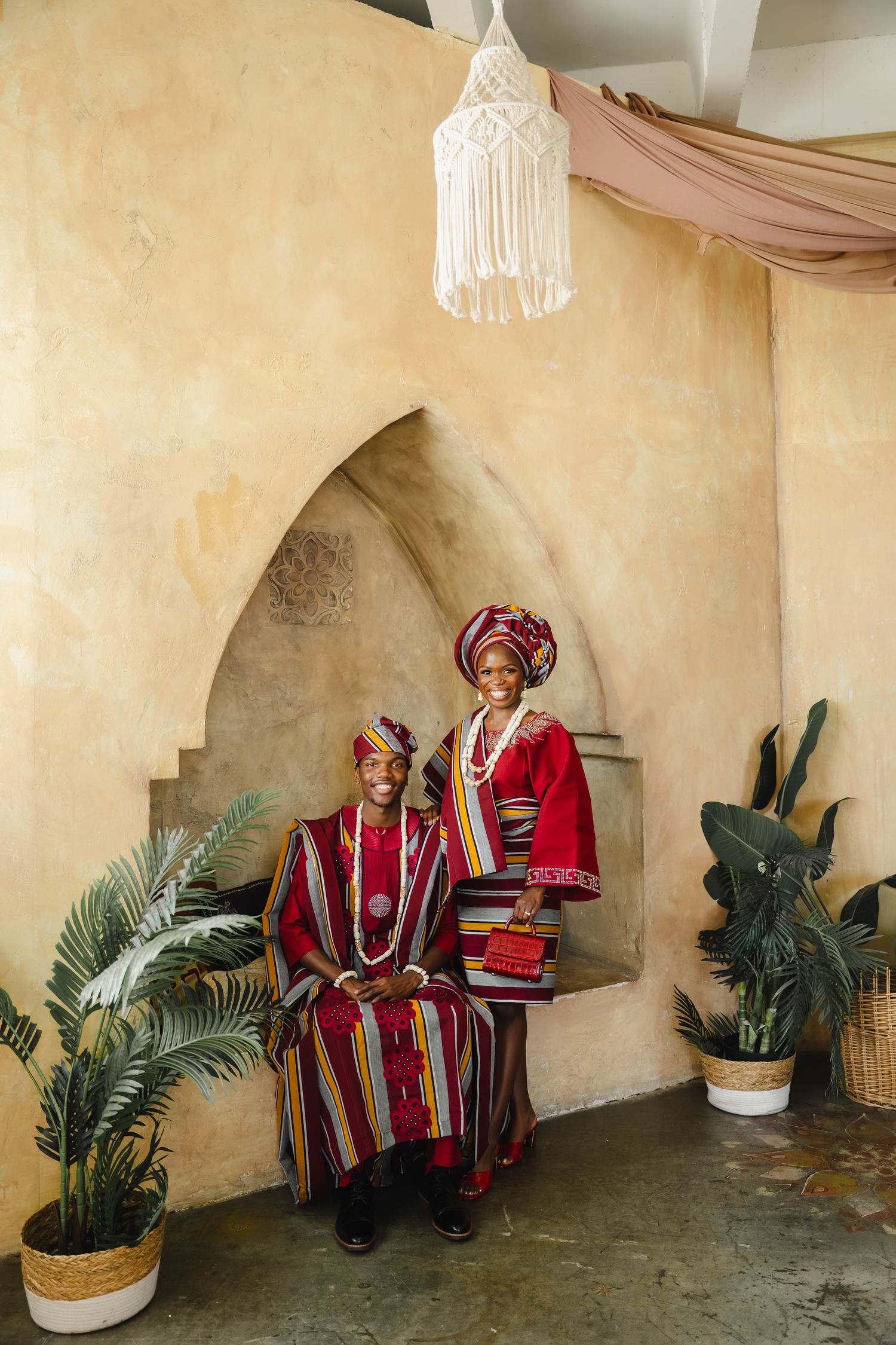Couple in vibrant traditional African attire posing together in a decorated venue with plants and macrame decor.