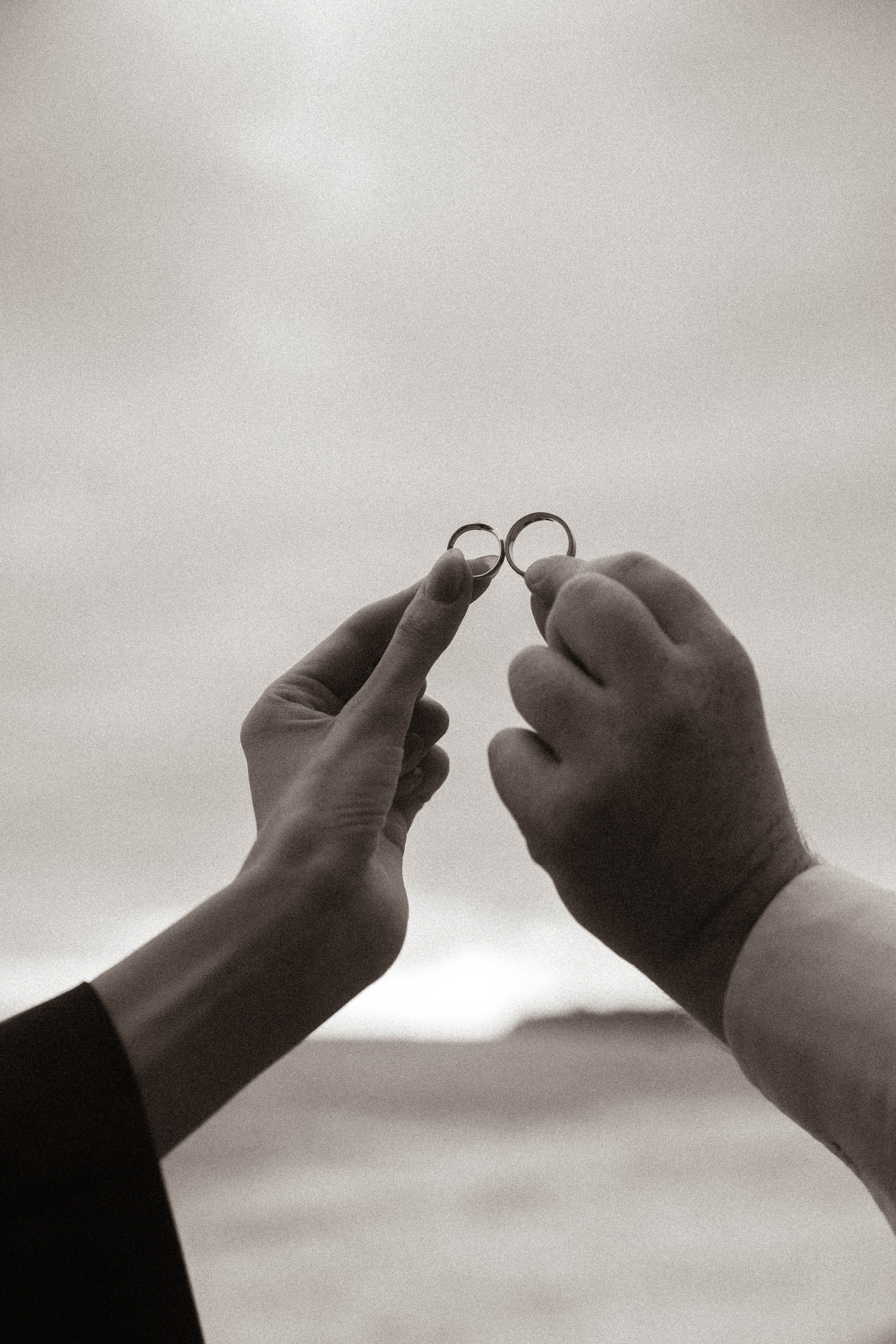 Two hands holding wedding rings up against a cloudy sky in a black and white photo.