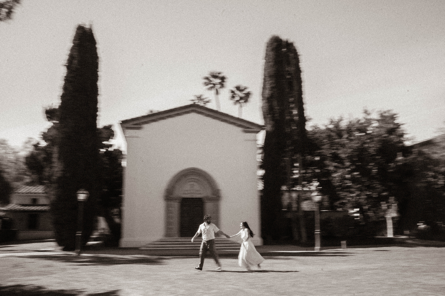 Black and white photo of a couple walking hand in hand past a white chapel with tall cypress trees.