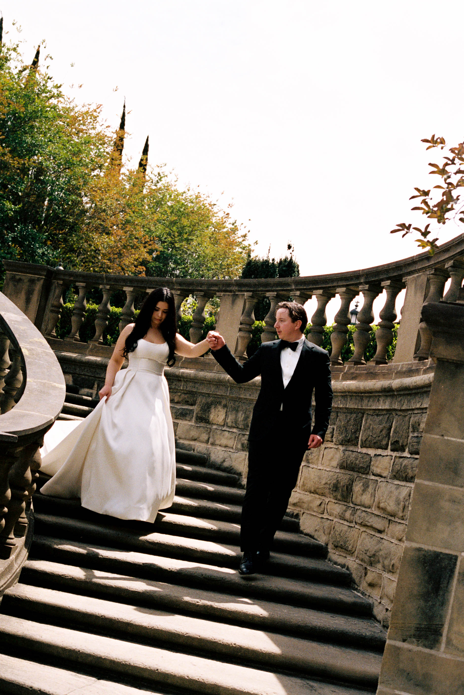 A bride and groom hold hands while walking up elegant stone stairs at an outdoor wedding venue surrounded by lush greenery.