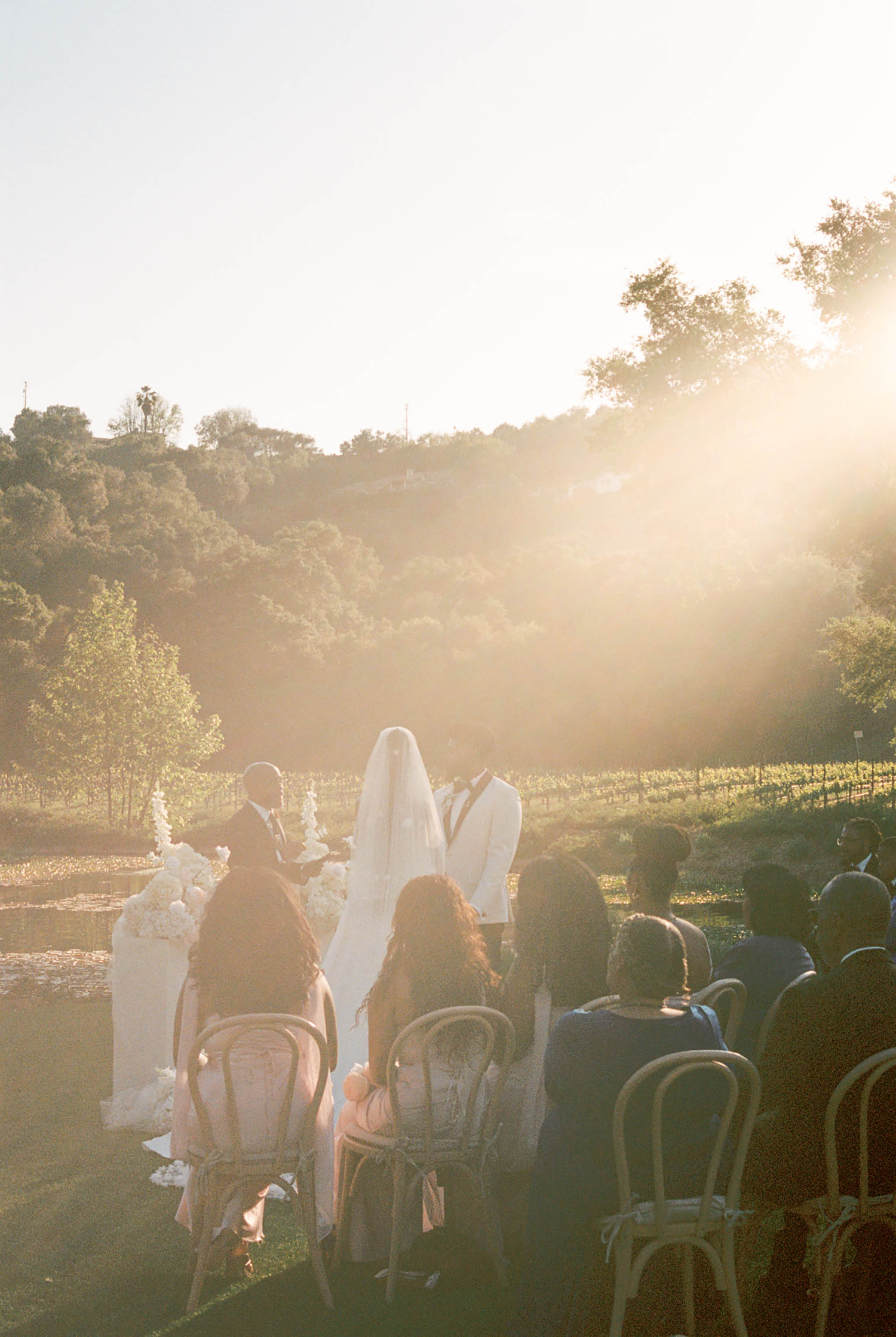 Outdoor wedding ceremony at golden hour with couple at altar, guests seated in wooden chairs, vineyard and hills backdrop.