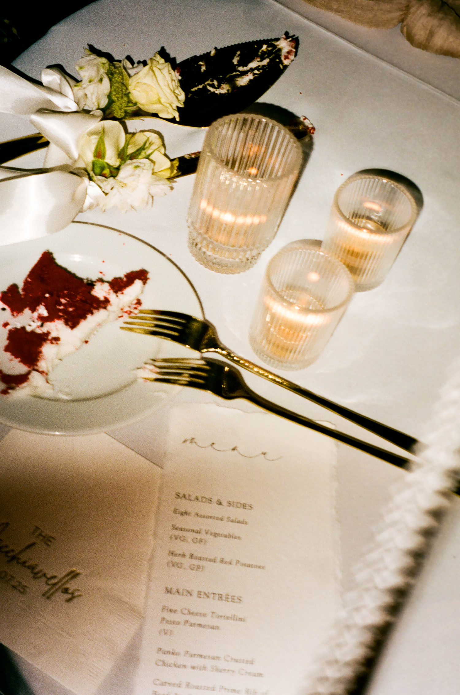 Elegant wedding reception table setting with red velvet cake slice, gold forks, glowing candles, and a dinner menu card.
