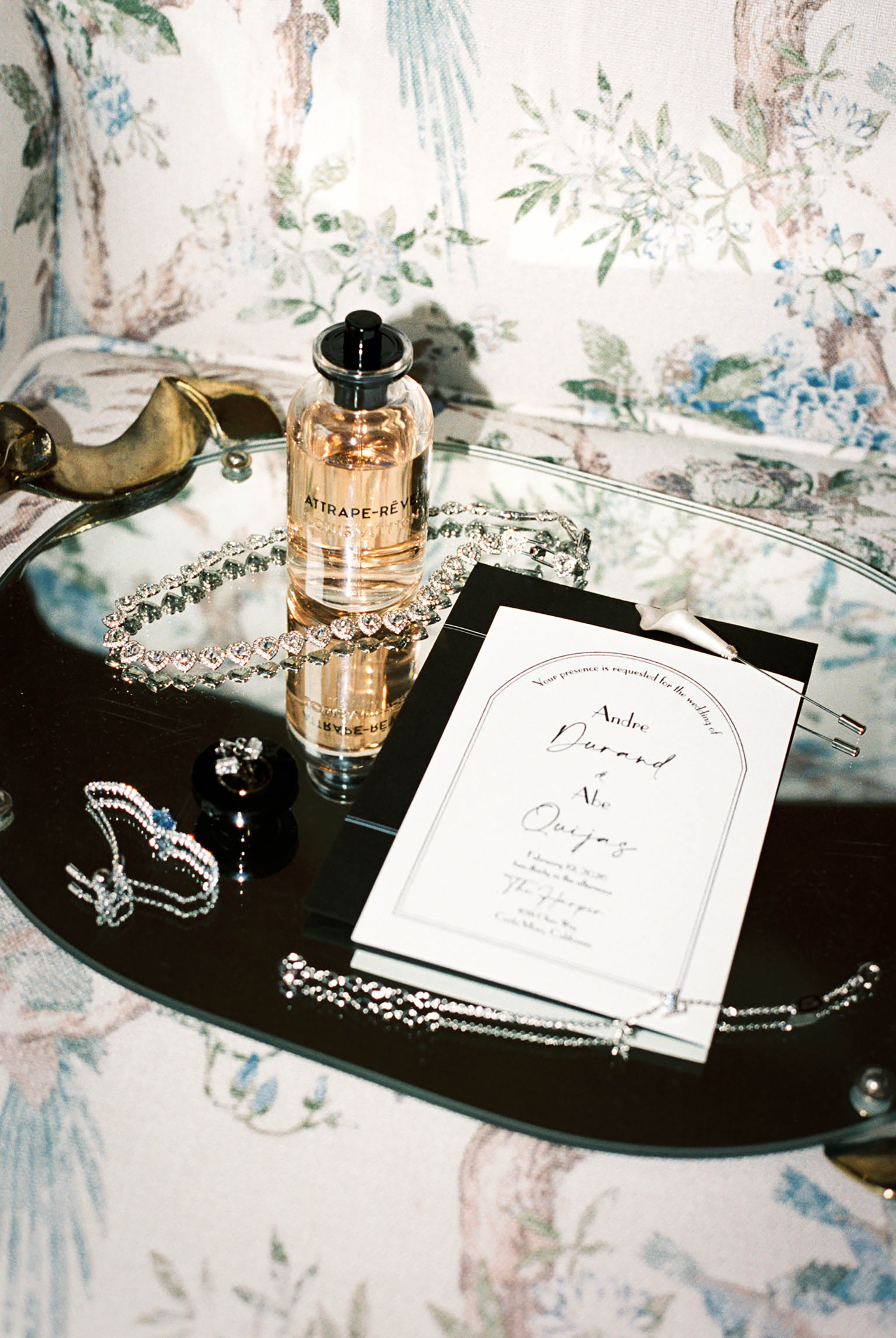 Elegant wedding invitation on mirrored tray with perfume bottle, crystal jewelry, and floral wallpaper backdrop.