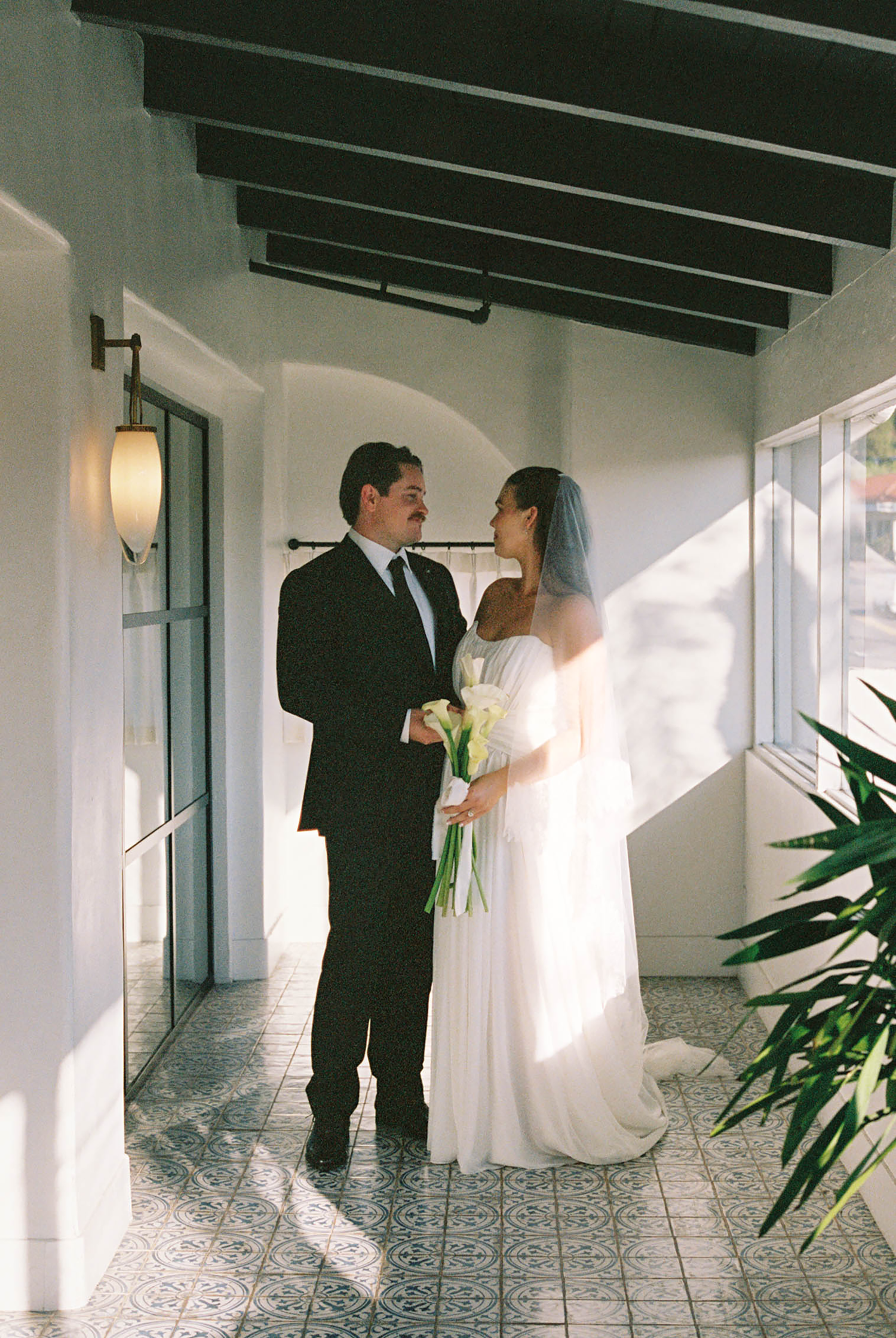 Bride and groom sharing a moment on sunlit tiled porch with calla lilies.