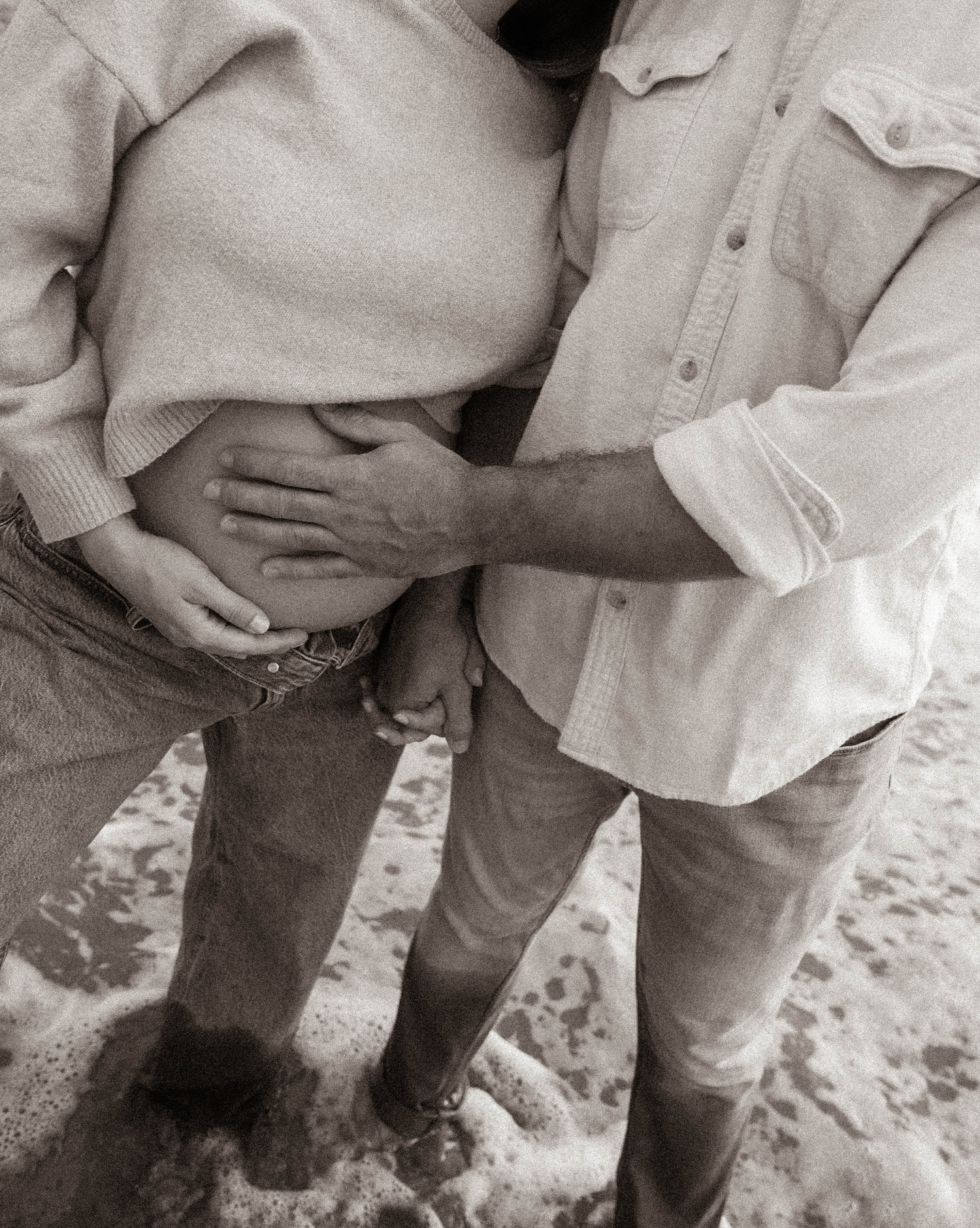Black and white close-up of couple holding hands on beach, man gently touching pregnant woman's belly.
