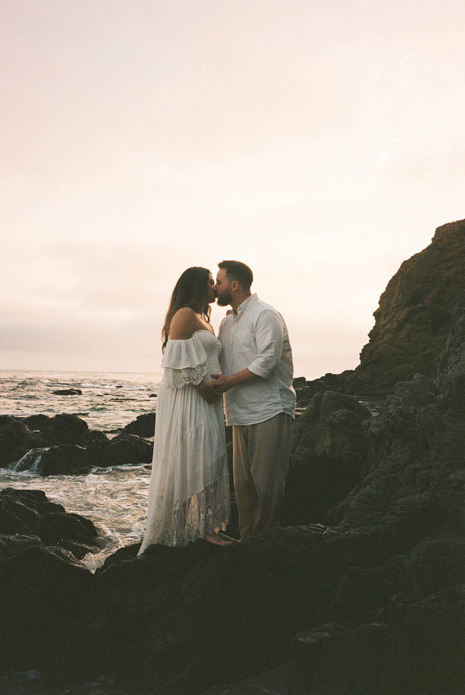 Expecting couple kissing near dark coastal rocks at sunset, maternity photography.