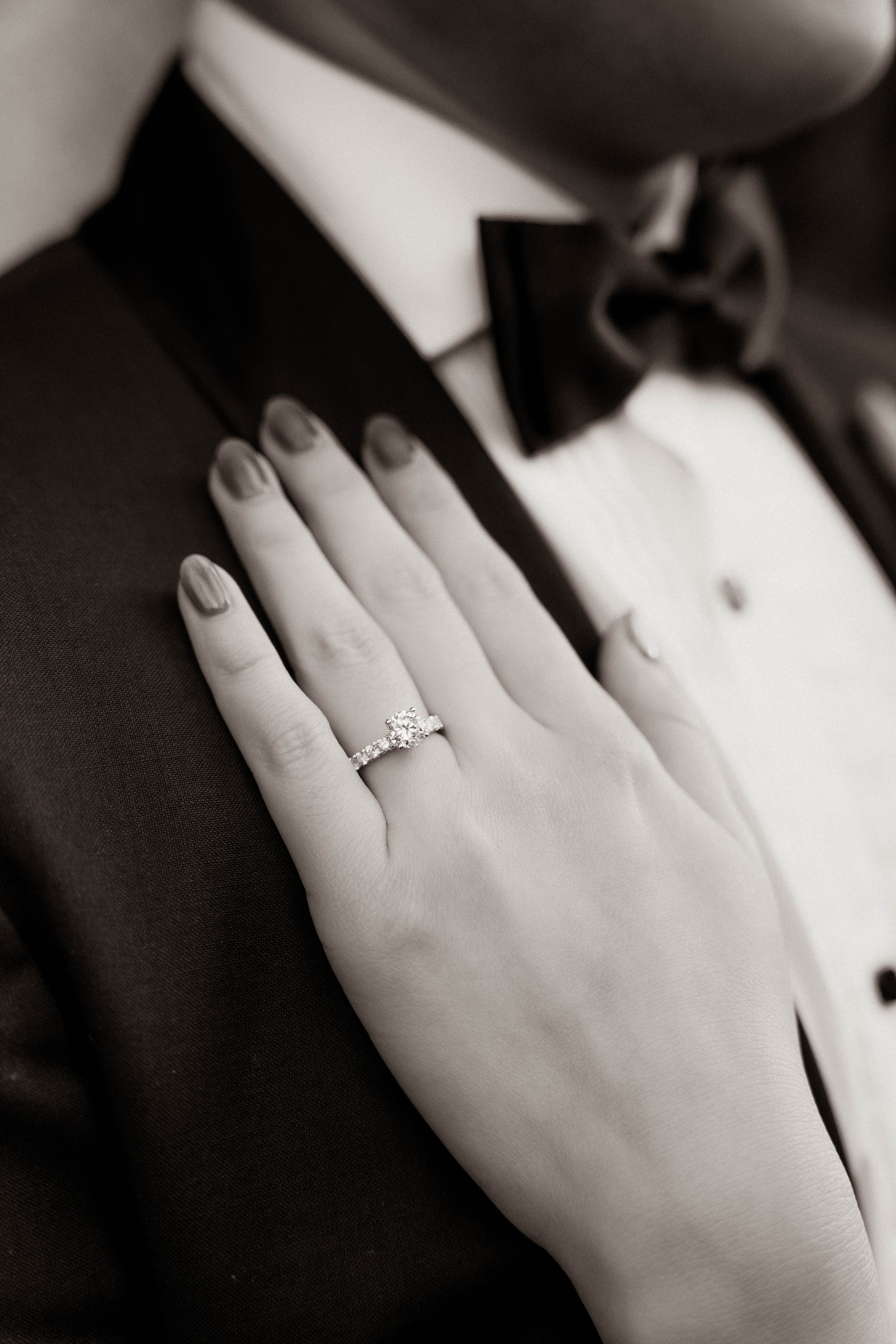 Black and white close-up of a diamond engagement ring on a bride's hand against a tuxedo.