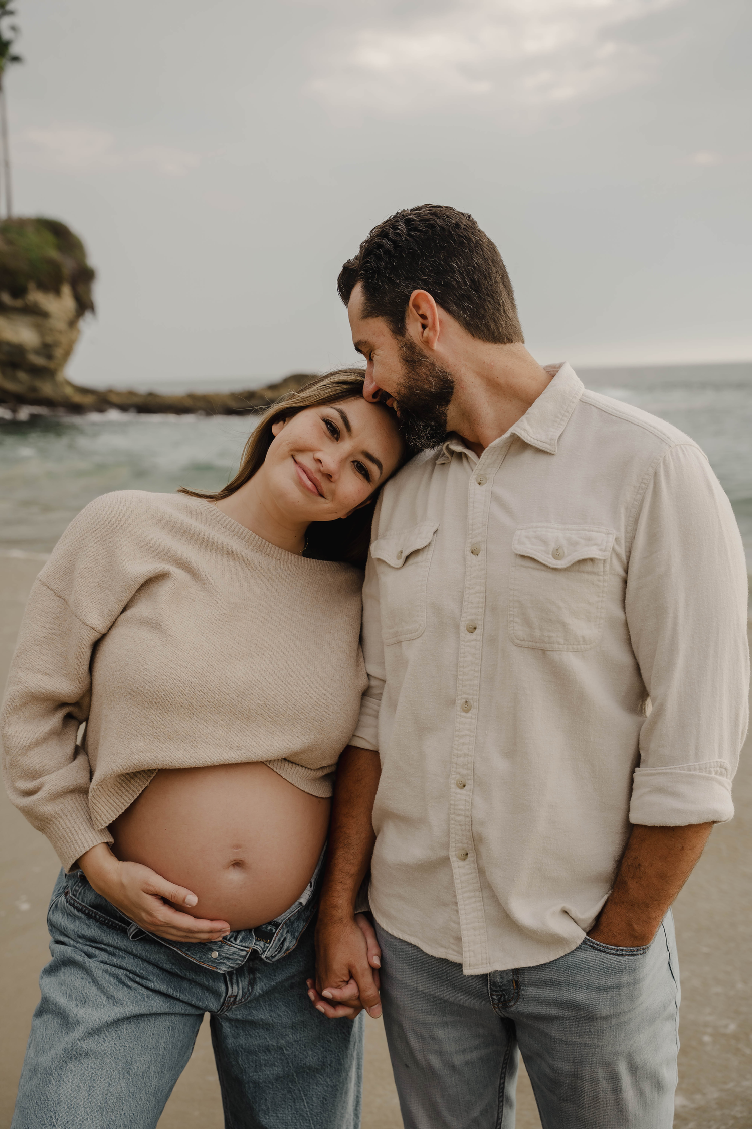 Pregnant couple embracing on a rocky beach, woman smiling with exposed baby bump, ocean backdrop.
